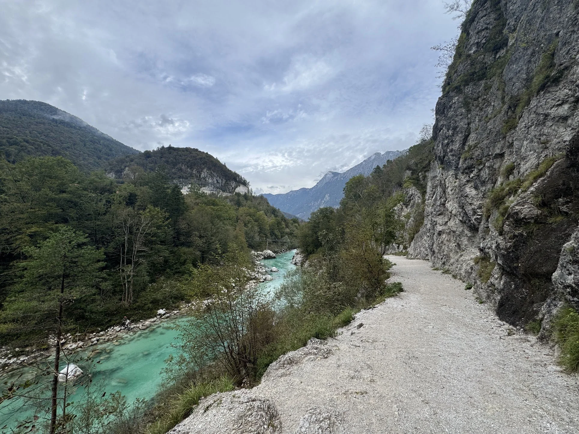 A scenic mountain landscape with a turquoise river flowing through a forested valley, a gravel hiking trail on the right, and rugged cliffs on the right side of the image, under a partly cloudy sky.