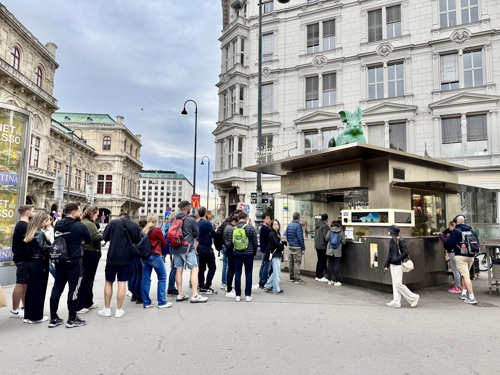 A crowd of people standing on a city street near a kiosk with a green rabbit sculpture on top. The background features ornate historic buildings with intricate architectural details.