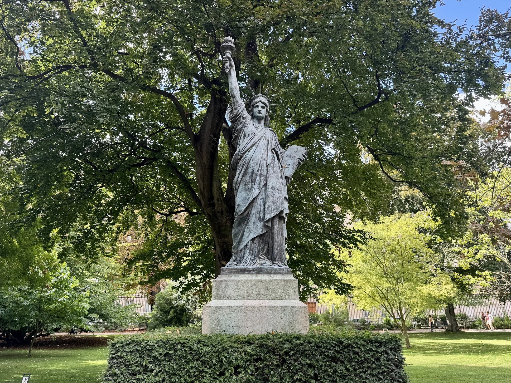 Statue of the Statue of Liberty, depicting a woman in robes holding a torch in her right hand and a tablet in her left, situated on a stone pedestal, surrounded by trees and greenery.