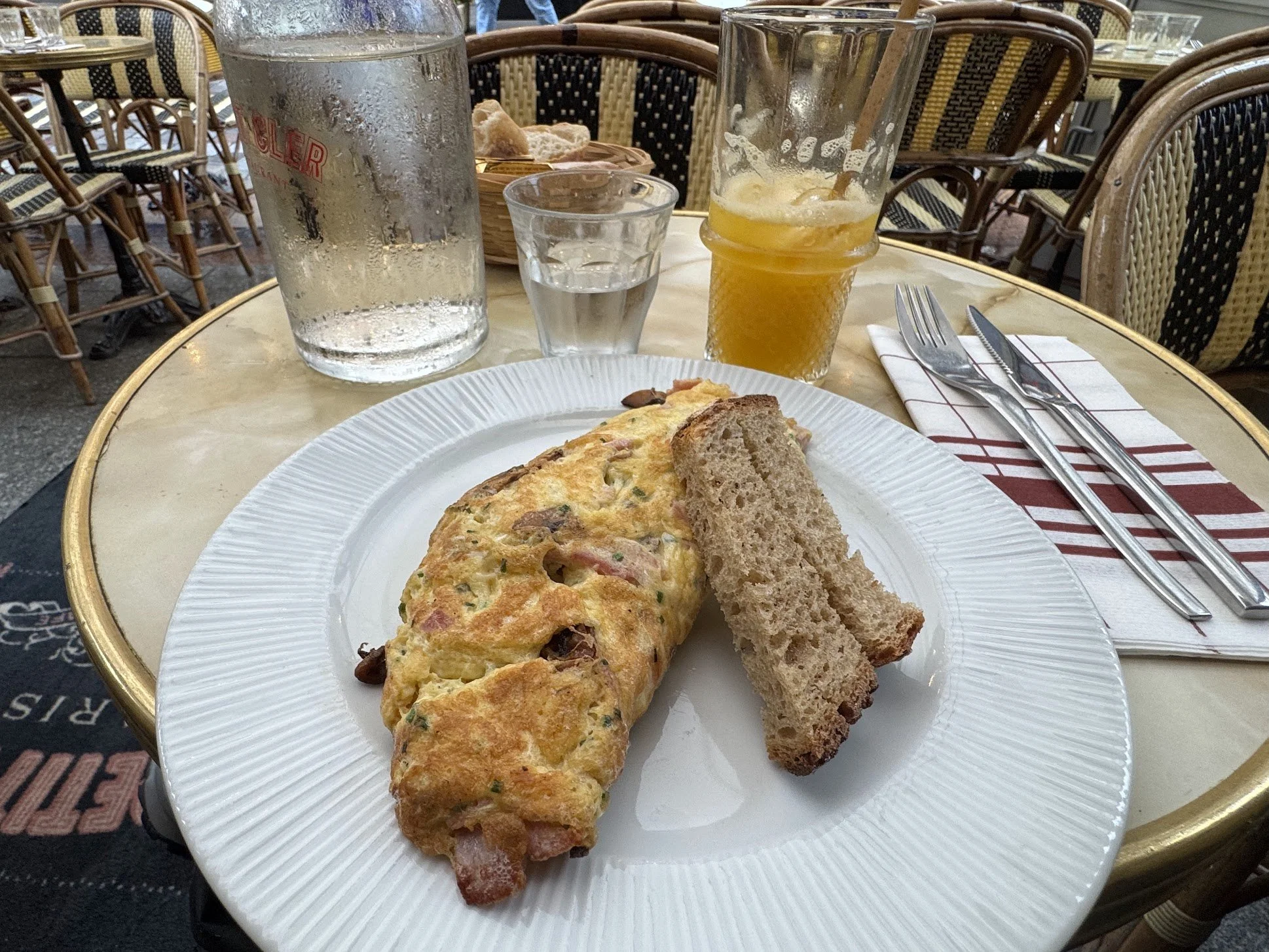 A breakfast table with a plate containing a veggie omelette and two slices of toasted bread. In the background, there is a large glass of water, a half-filled glass of orange juice, and a pitcher of water. The table is outdoors, surrounded by wicker 