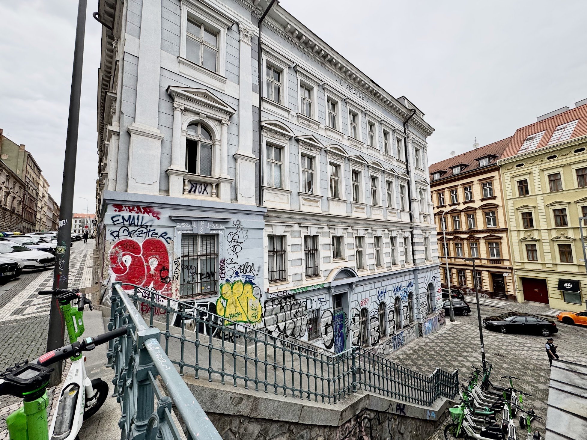 A multi-story historical building with white and light blue walls, decorative architectural features, and graffiti on the lower part, situated on a city street with parked cars, a bicycle, and neighboring buildings with reddish and beige facades.