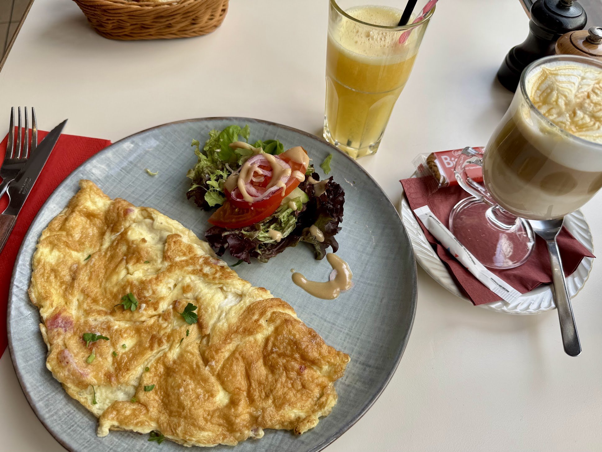 A breakfast plate with a cheese omelette, a side salad with lettuce, tomato, onion, and dressing, a tall glass of orange juice, and a glass of coffee topped with whipped cream, on a white table with a red napkin, utensils, and condiments.