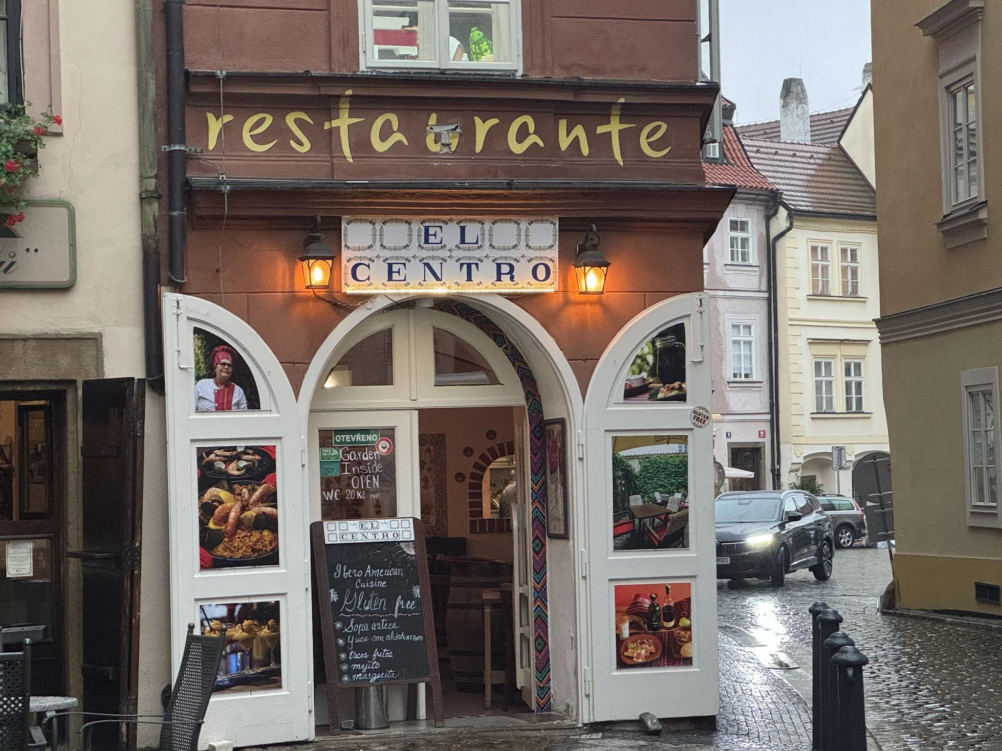 A small restaurant on a wet street with a sign reading 'EL CENTRO.' The storefront has images of food, including dishes with meat and vegetables. A chalkboard menu is inside, listing items such as Mexican cuisine, gluten-free options, and various dishes like tacos and mojitos. The building has decorative elements and warm lighting, and there are cars parked on the street outside.