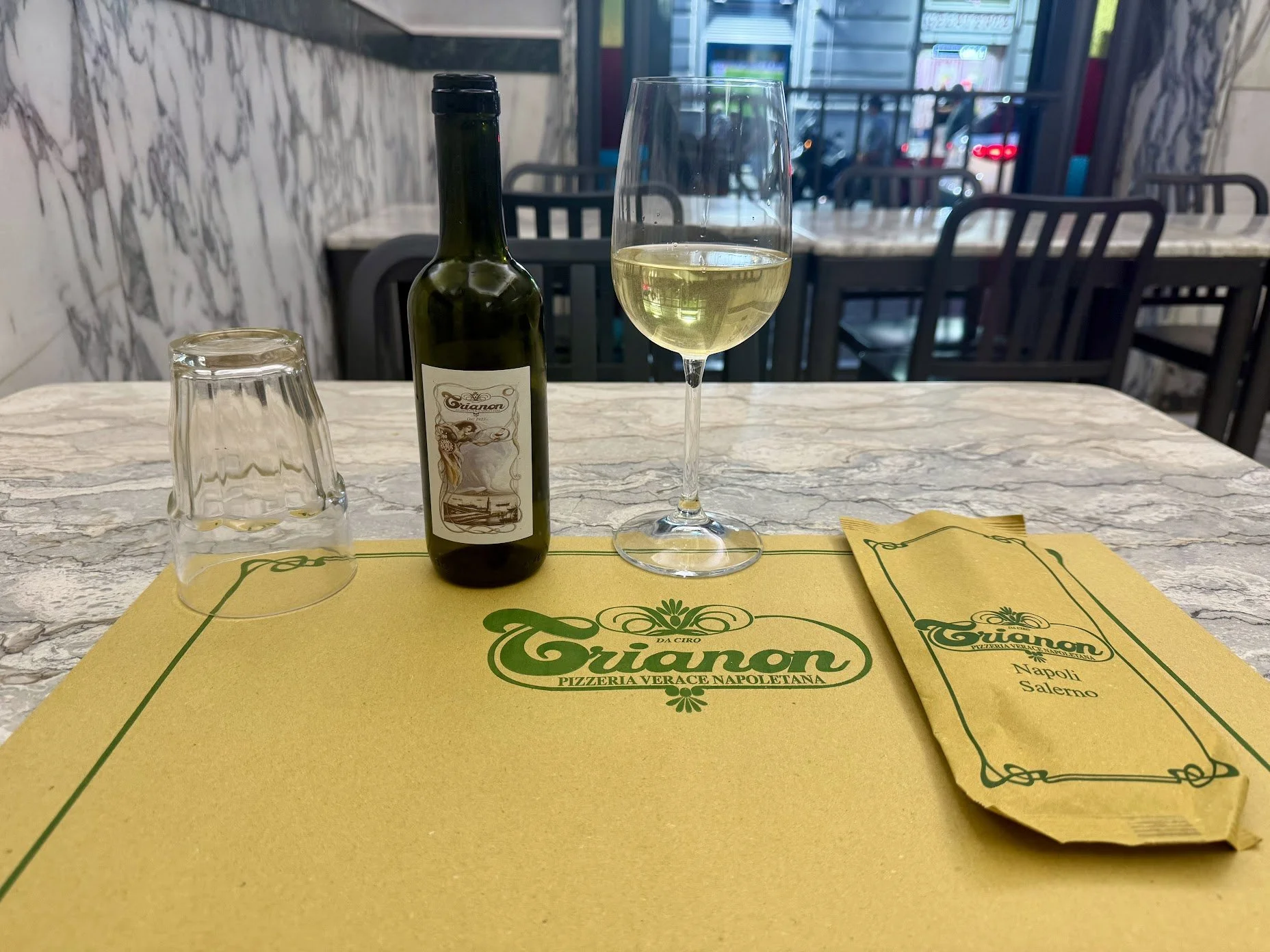 A glass of white wine, a dark green wine bottle with a vintage-style label, an upside-down glass, and a yellow napkin with green text that reads "Grianon Pizzeria Verace Napoletana" on a marble table. In the background, there are chairs and windows o
