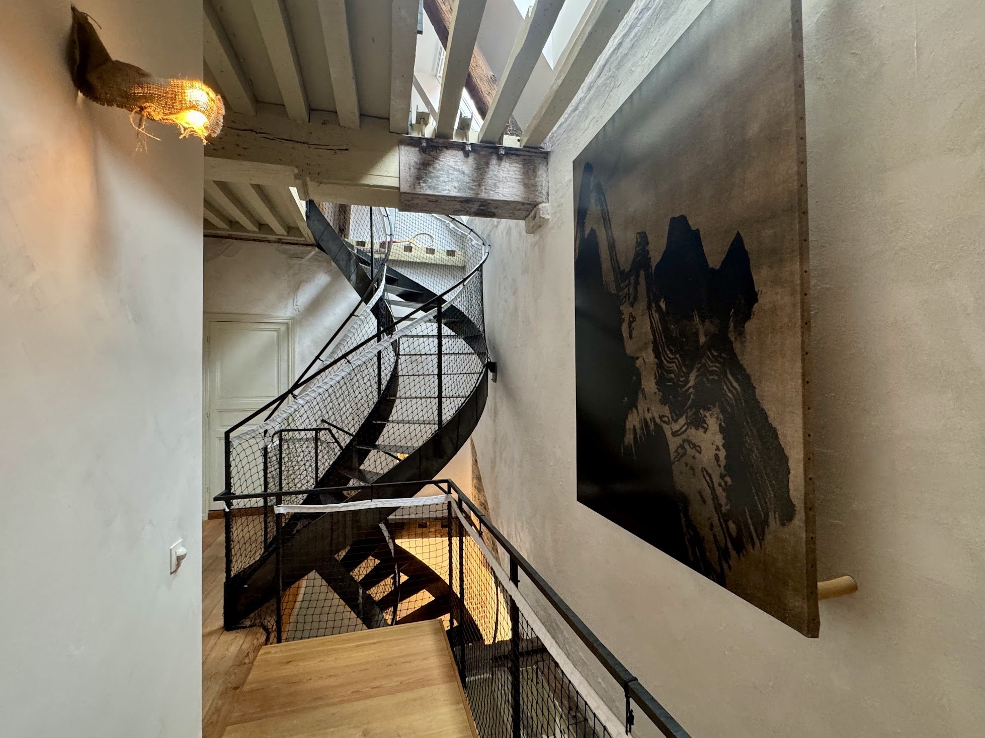 Interior view of a modern house showcasing a black metal spiral staircase with wooden steps, a large rectangular black and brown abstract mountain painting on a beige wall, exposed wooden beams on the ceiling, and a concrete floor near a window that 