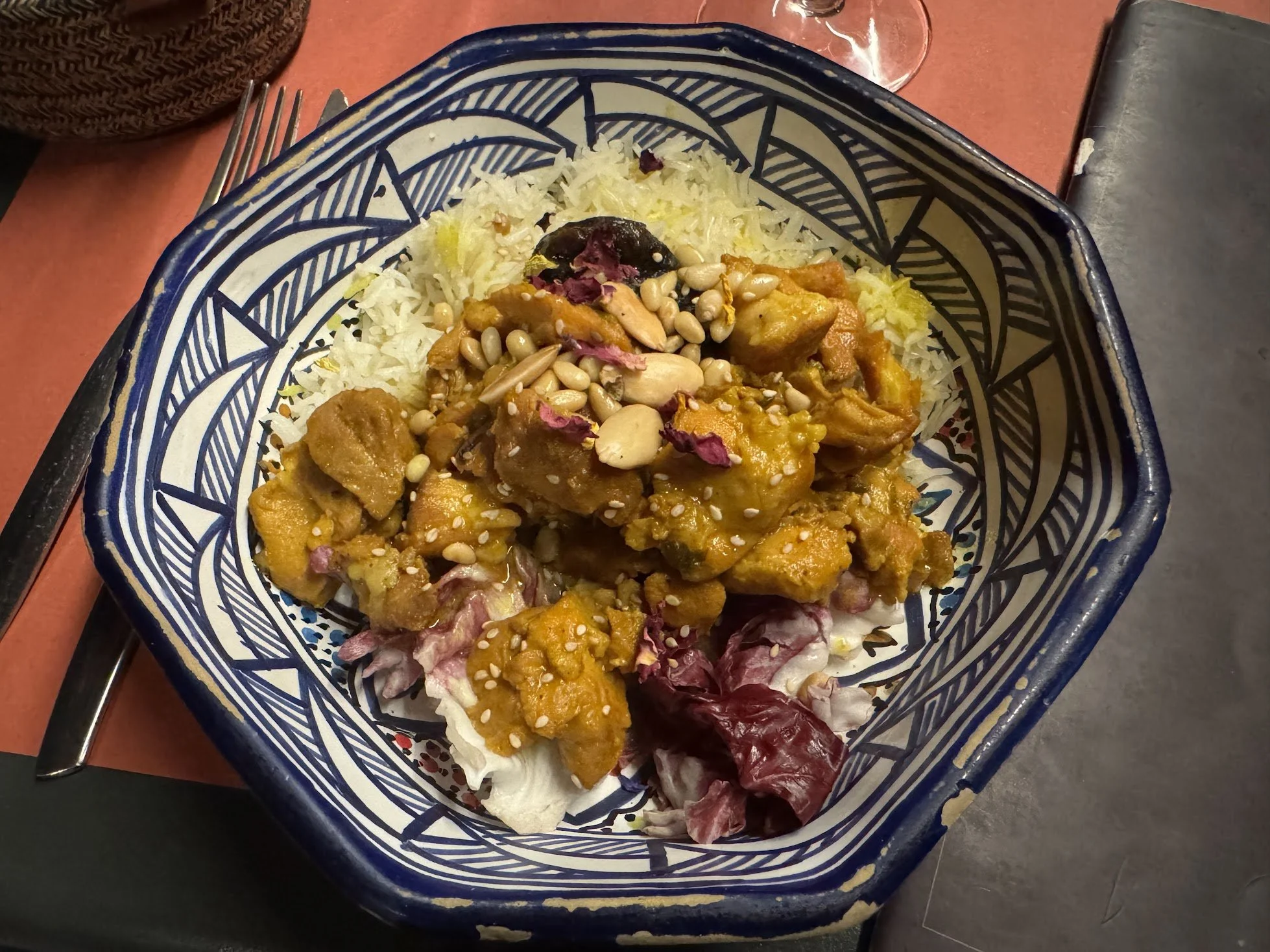 A bowl of rice topped with a curry, garnished with pine nuts and purple flower petals, placed on a table with a fork and a glass in the background.