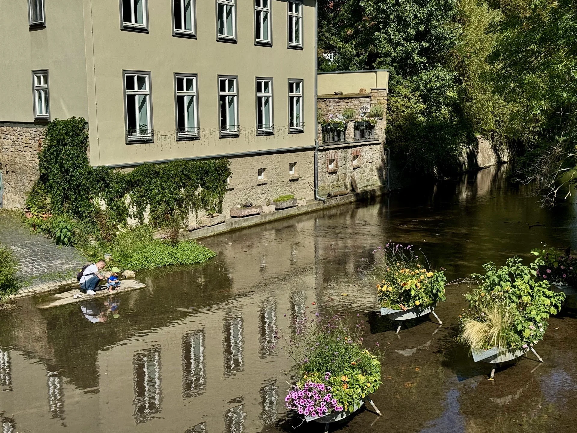 Two people, an adult and a child, sitting by a river's edge with colorful flowers in planters nearby. A building with multiple windows and a stone wall line the riverbank, with lush trees in the background.