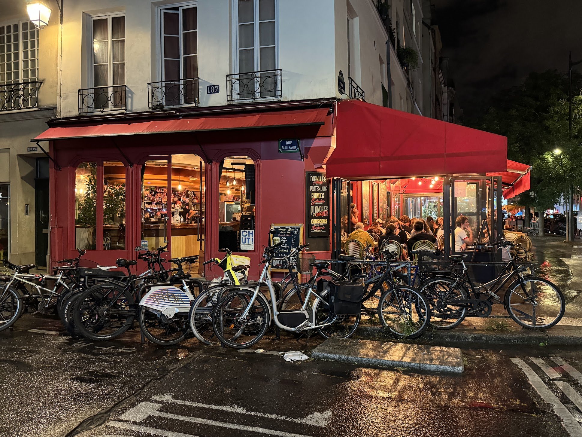 A street scene at night show a red restaurant or cafe with large windows and an outdoor seating area filled with people. The street has many parked bicycles in the foreground, and the street is wet, likely from rain. There are buildings with windows 