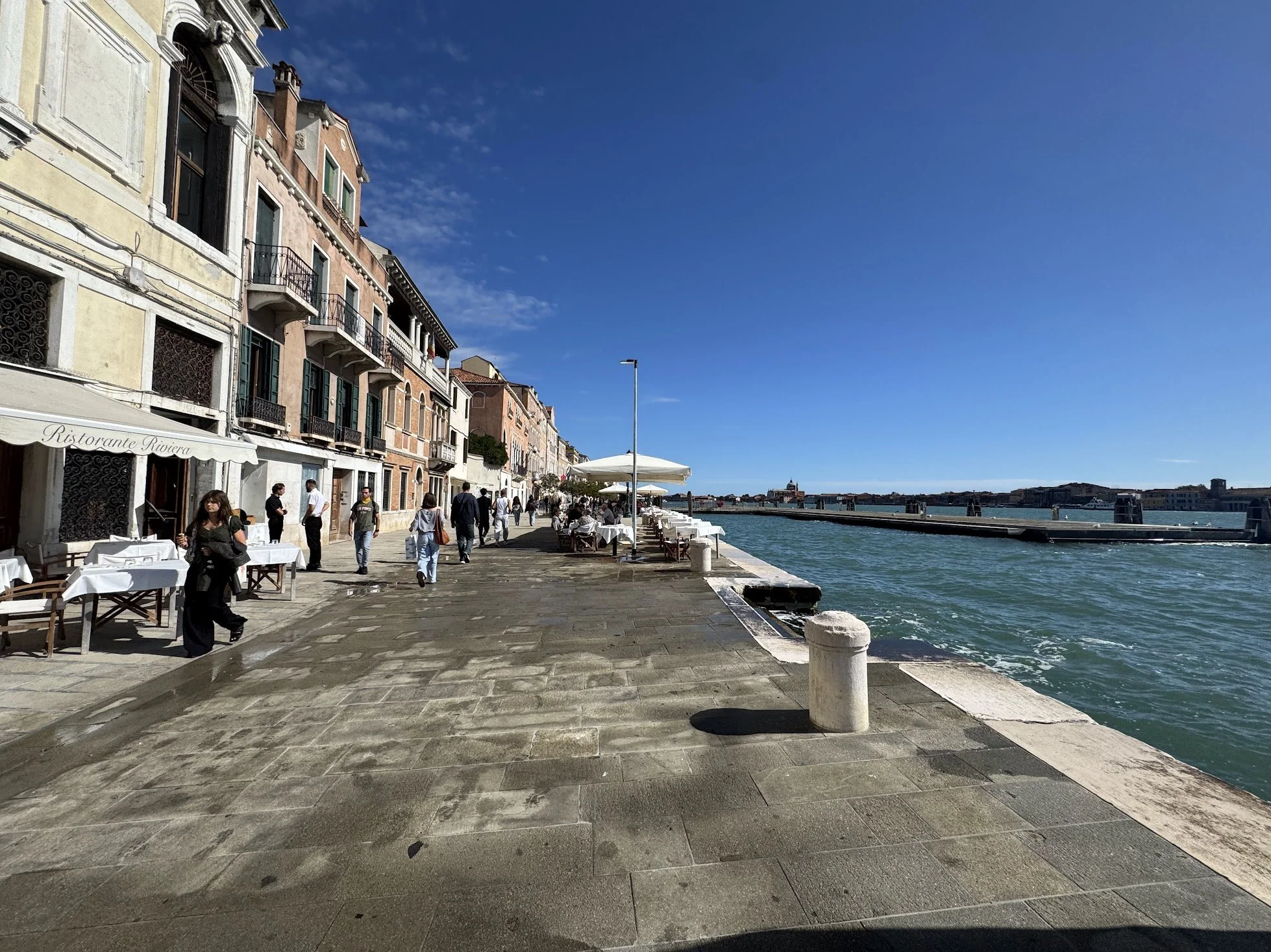 View of a waterfront promenade with outdoor dining tables, colorful Italian-style buildings, and a clear blue sky.