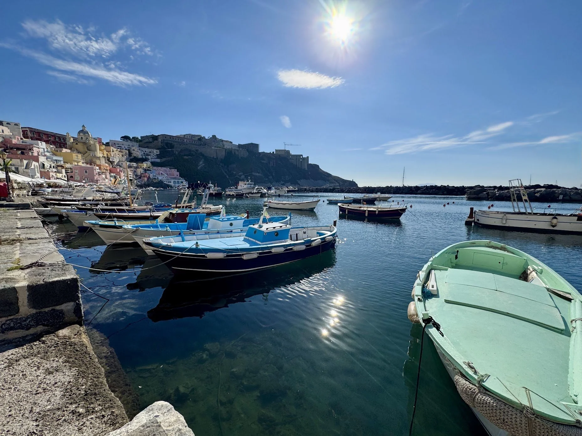 A harbor with several small boats docked along the stone quay, overlooking a hillside city with colorful buildings, under a clear blue sky, with the sun shining brightly.