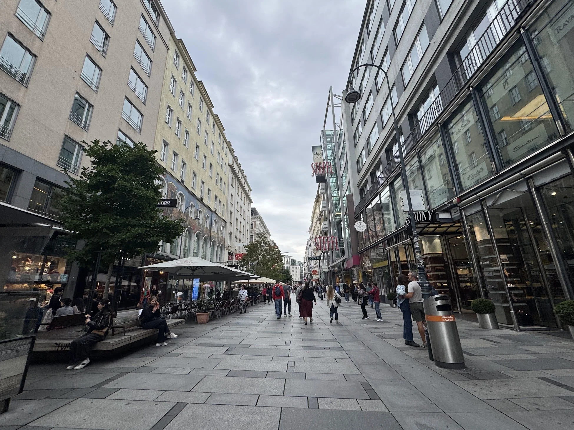 A busy city street filled with pedestrians, outdoor seating, and various storefronts, with tall buildings on both sides and a cloudy sky overhead.