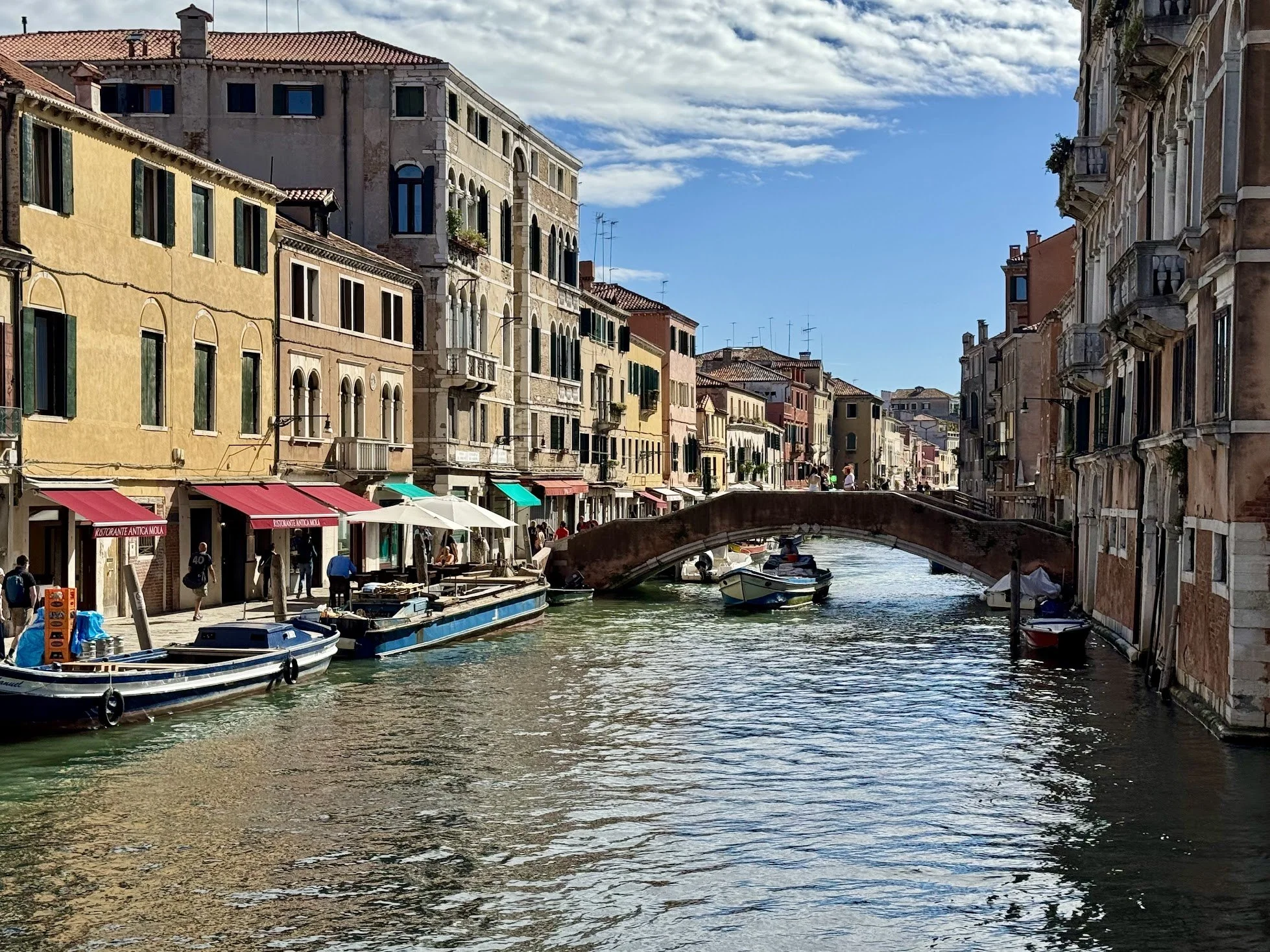 View of Venice canal with boats, historic buildings, pedestrians, and a stone bridge under a partly cloudy sky.