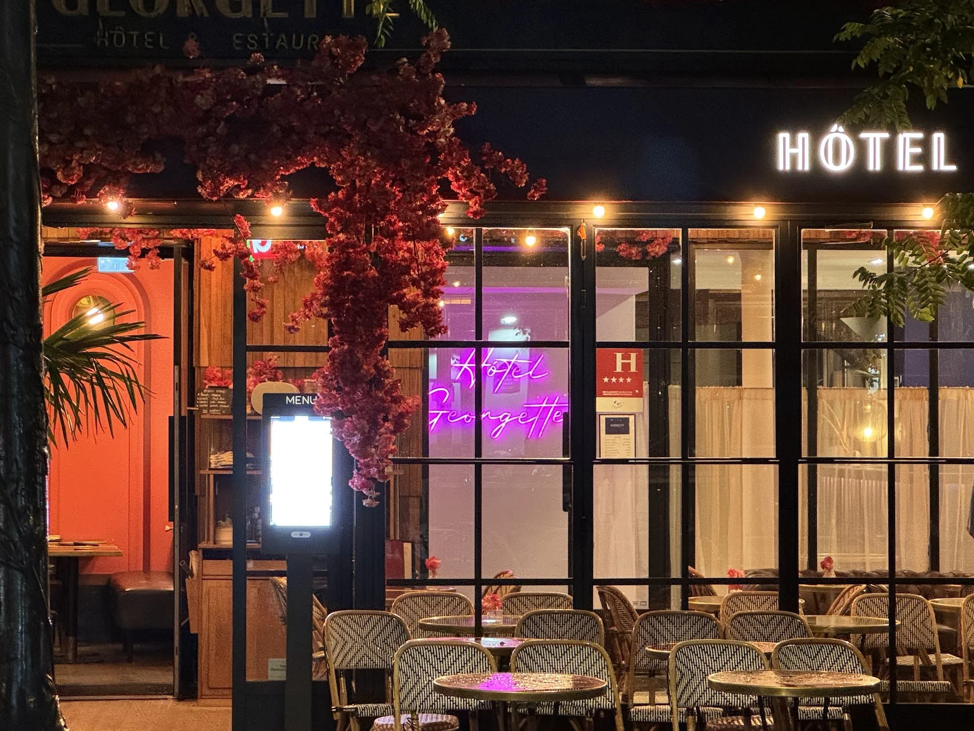 Nighttime view of a restaurant's interior and exterior through large glass windows, with neon signs, artificial flowers, and empty chairs.