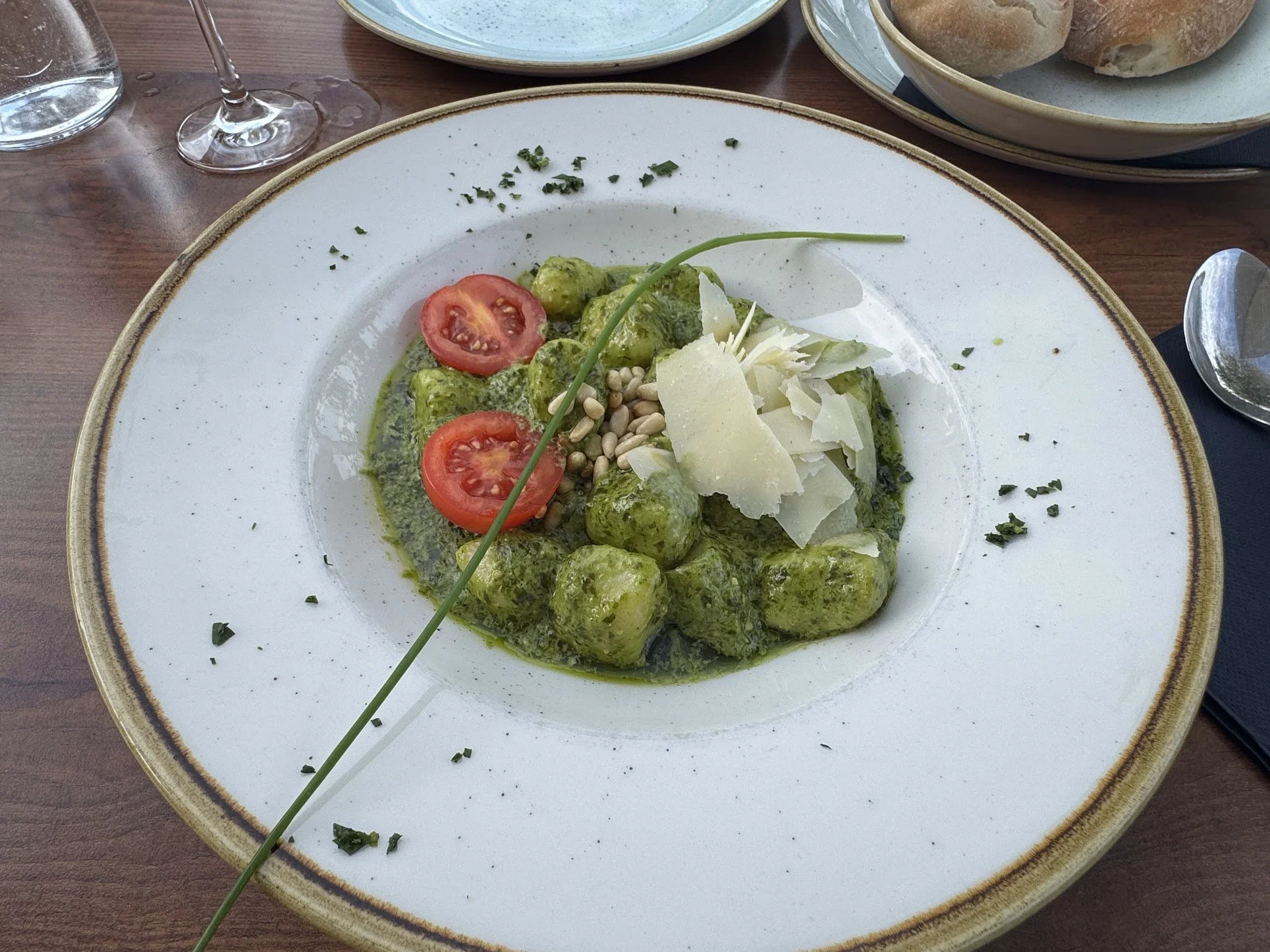 A white plate with green gnocchi, cherry tomato slices, pine nuts, and shredded cheese, garnished with chopped herbs and a chive on top. A drink glass and bread are also visible on the wooden table.