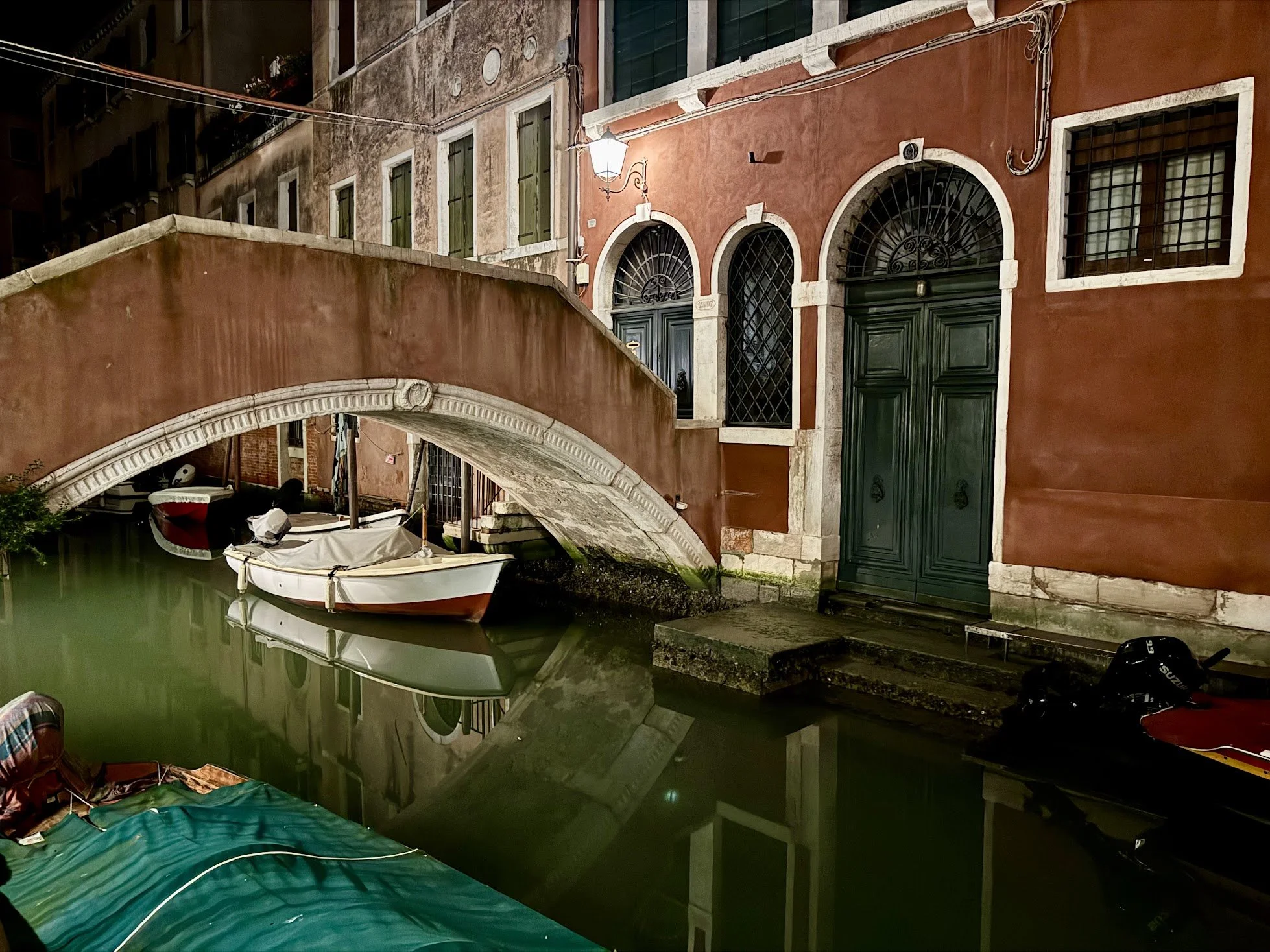 Nighttime view of a canal in Venice, Italy, with boats docked and old buildings with barred windows and green doors reflecting on the water.