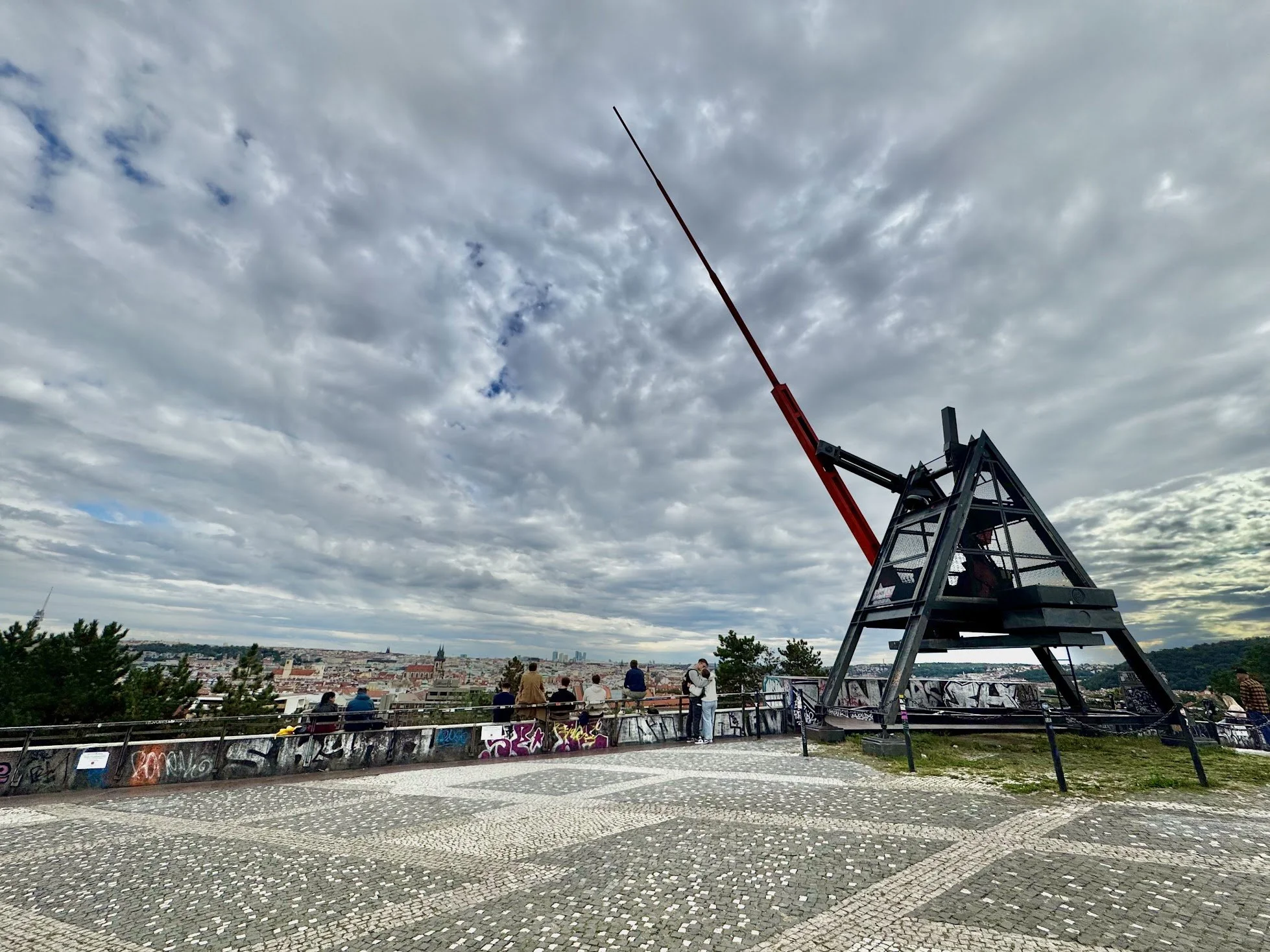 People sitting and standing on an observation platform with a view of a city skyline and a panoramic sky with clouds, and a large crane structure in the foreground.