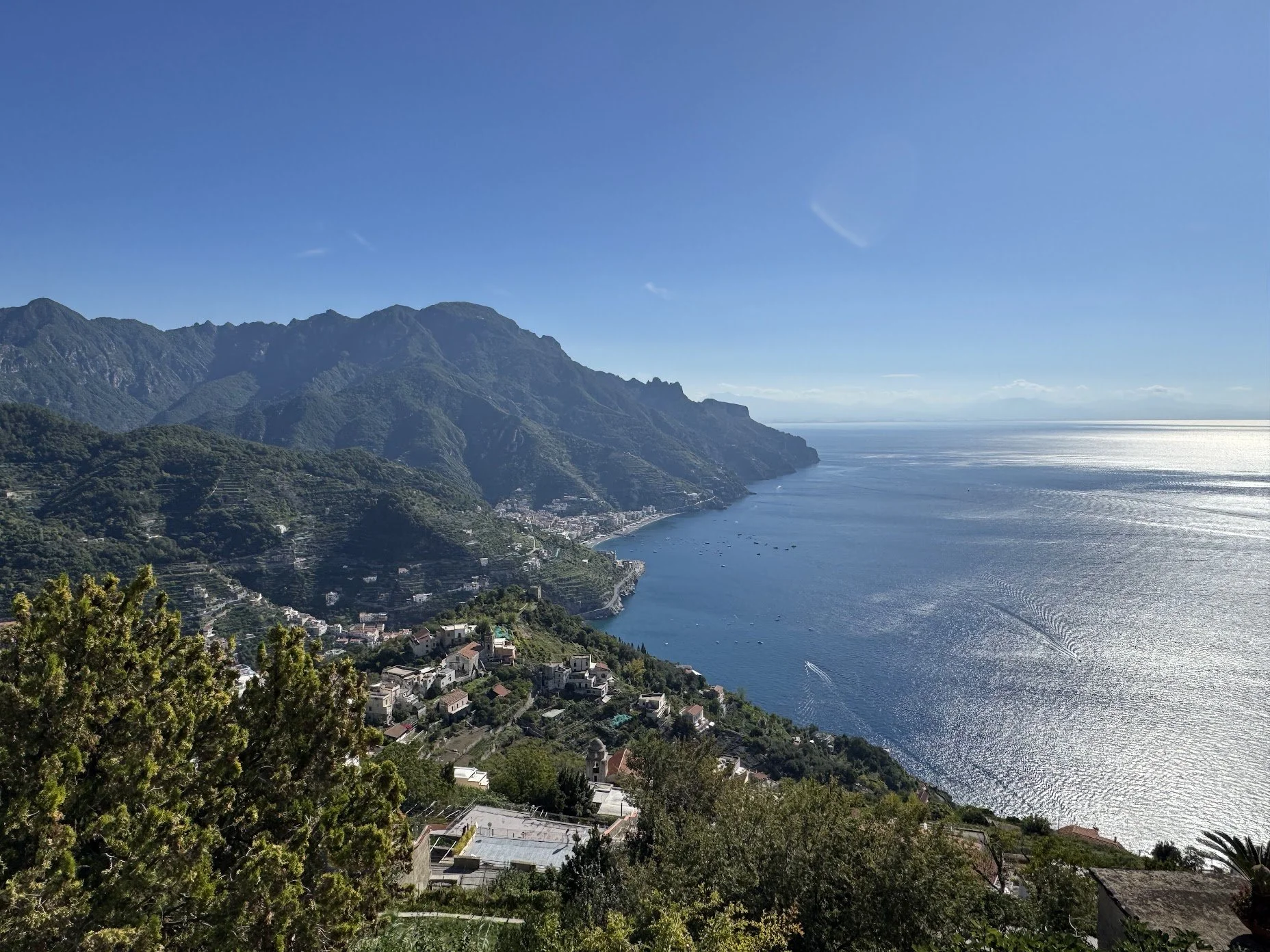 Coastal view of green mountain hills with a small town along the shoreline and boats on a blue ocean, under a clear sky.