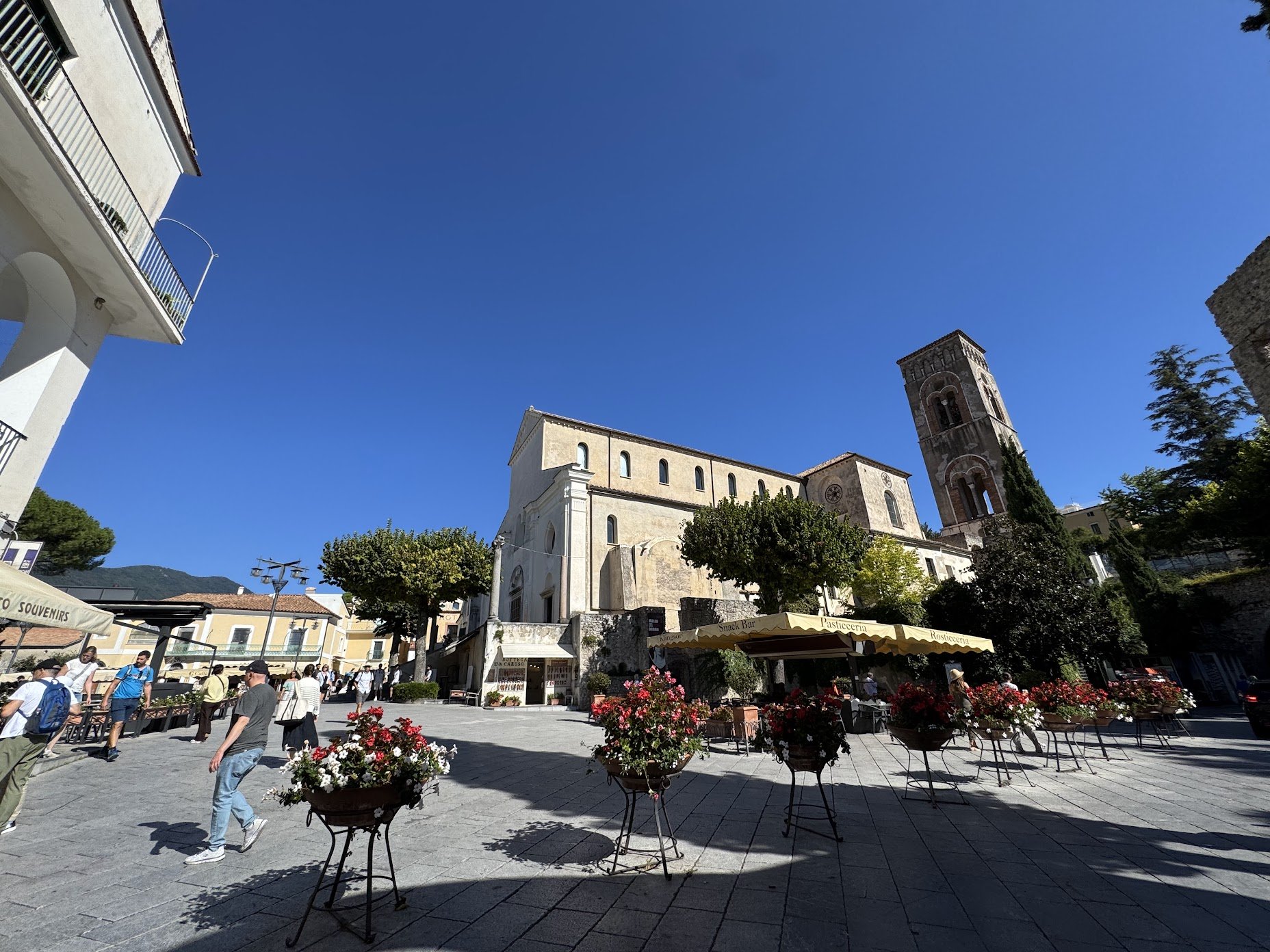 A lively outdoor plaza with people walking and sitting at tables, surrounded by colorful flowers in pots. In the background, there is a historic church with a tall bell tower and a stone facade, under a clear blue sky, with several trees and a small 