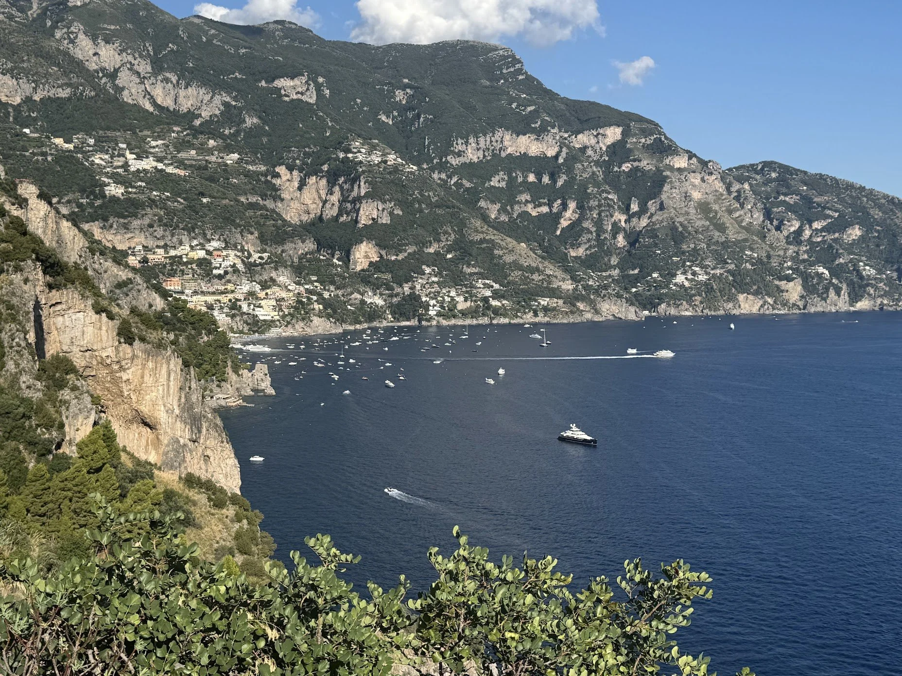 A coastal landscape featuring a large hillside with houses, rugged cliffs, and a deep blue sea with boats and yachts, under a partly cloudy sky.