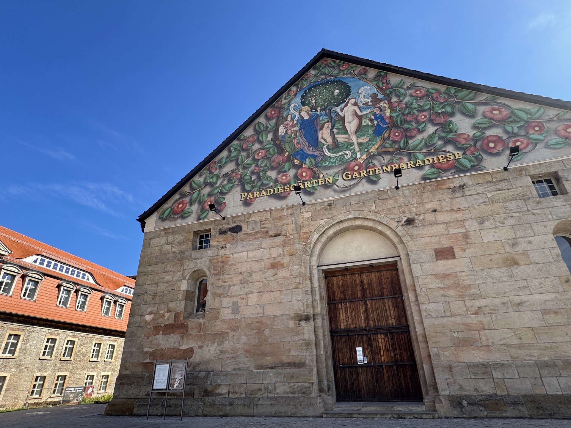 A stone church building with a large wooden door and a colorful mosaic depicting Adam and Eve in the Garden of Eden, surrounded by plants and flowers, on the front facade with the words "Paradies Gärten Gartenparadiese".