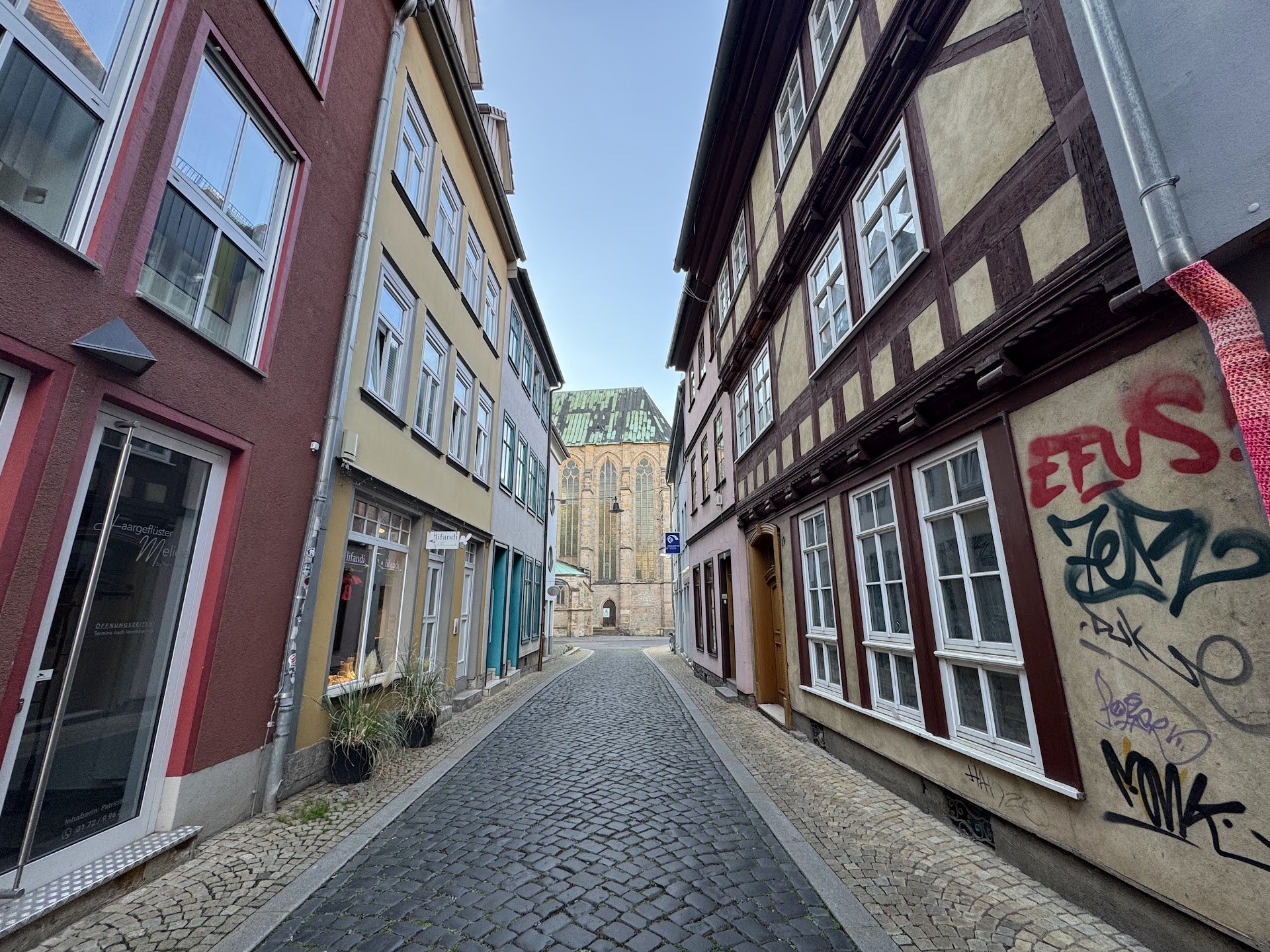 Narrow cobblestone street lined with colorful buildings on both sides, leading to a church in the background with a green, slightly weathered roof. One building has graffiti on its wall.
