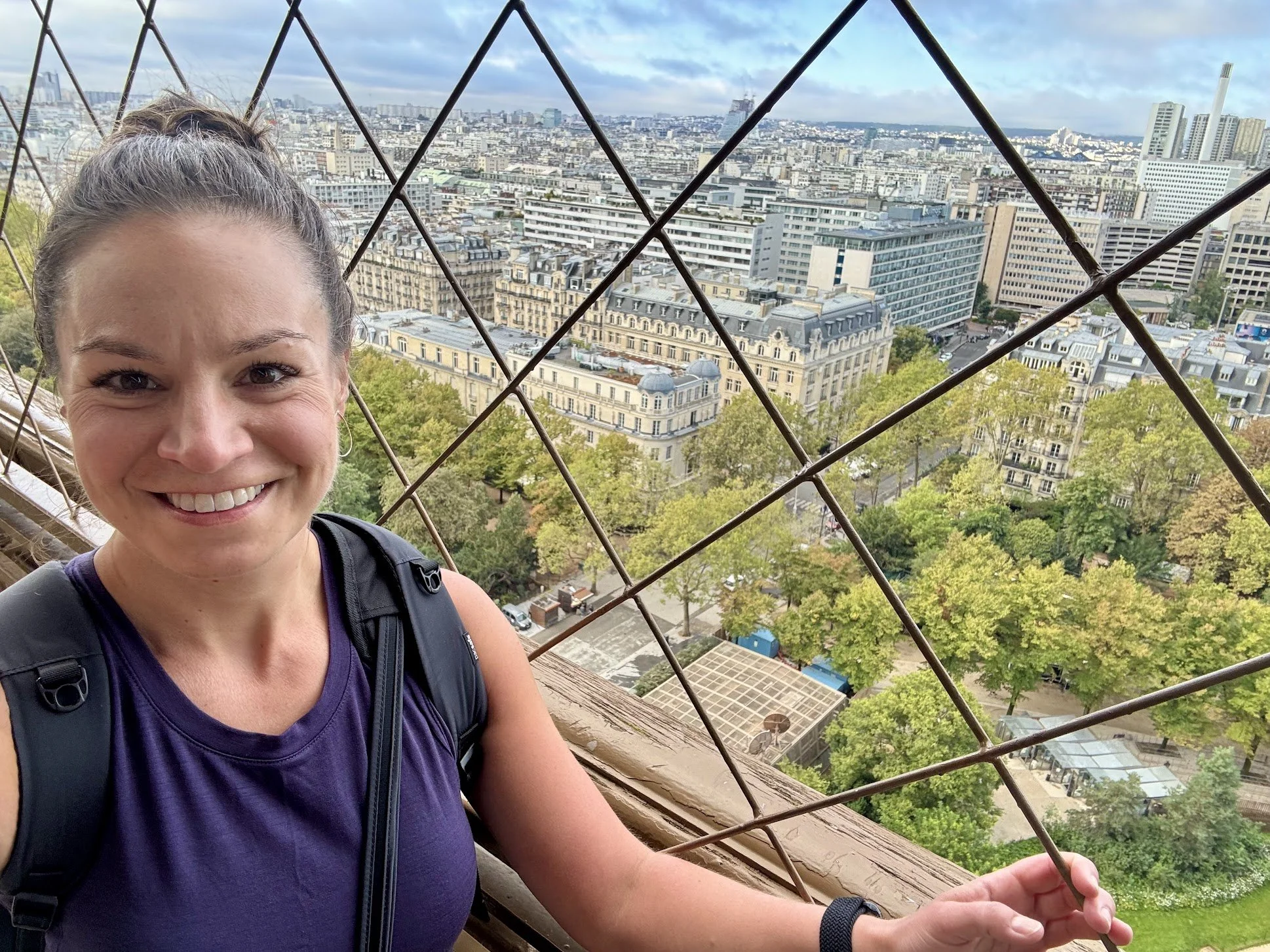 A smiling woman with a ponytail, wearing a purple tank top and a black backpack, standing on an outdoor observation deck with a metal safety fence, overlooking a cityscape with buildings, trees, and a cloudy sky.
