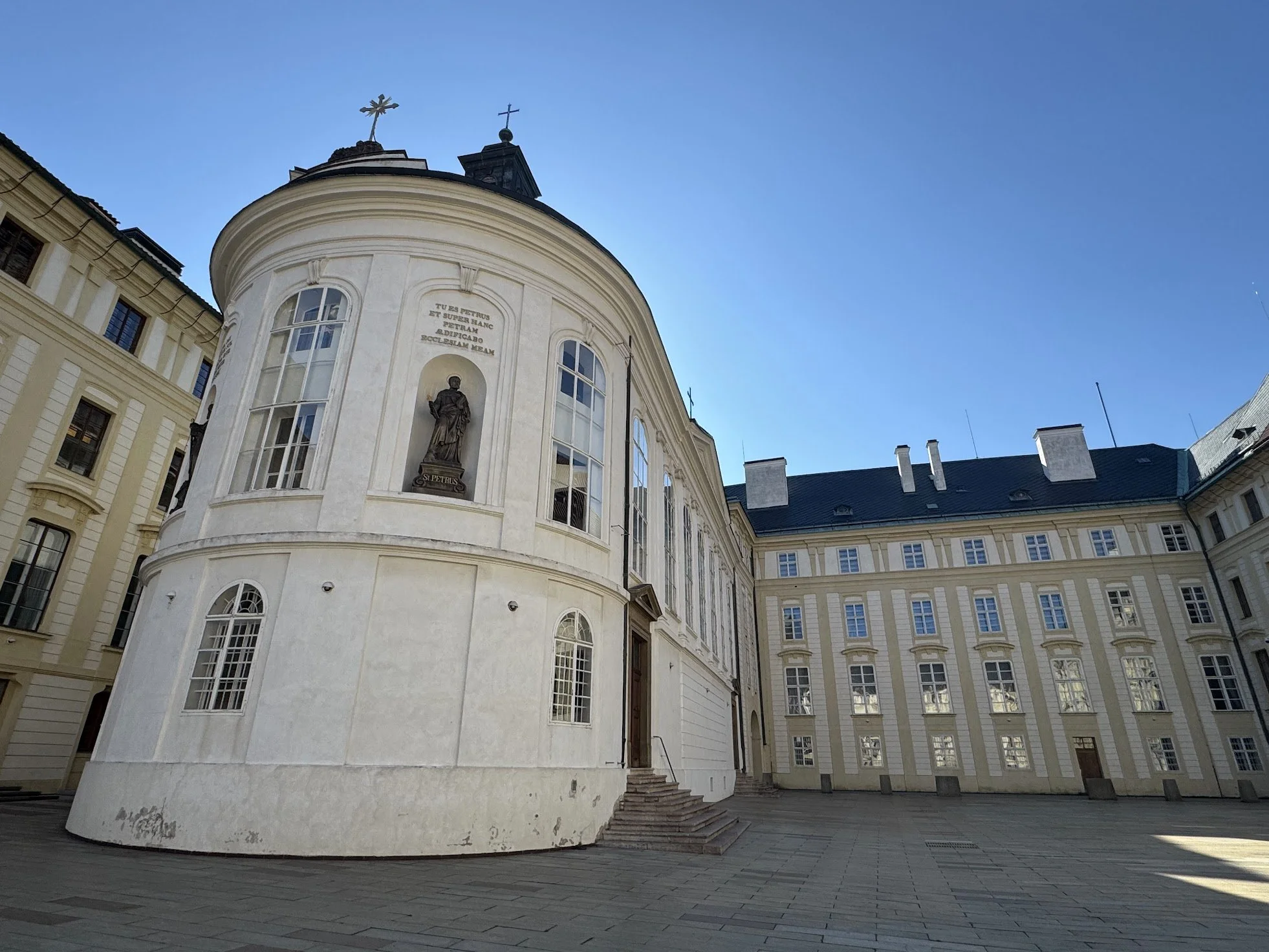 A church with a rounded facade and statues, surrounded by an elegant cream-colored building with multiple windows under a clear blue sky.