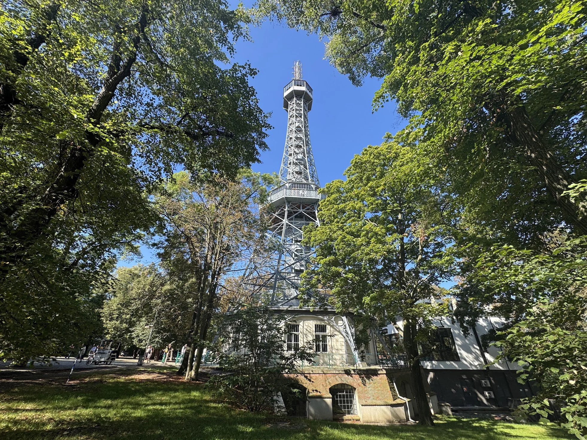 A tall, metallic television tower viewed through trees with green leaves and a clear blue sky.