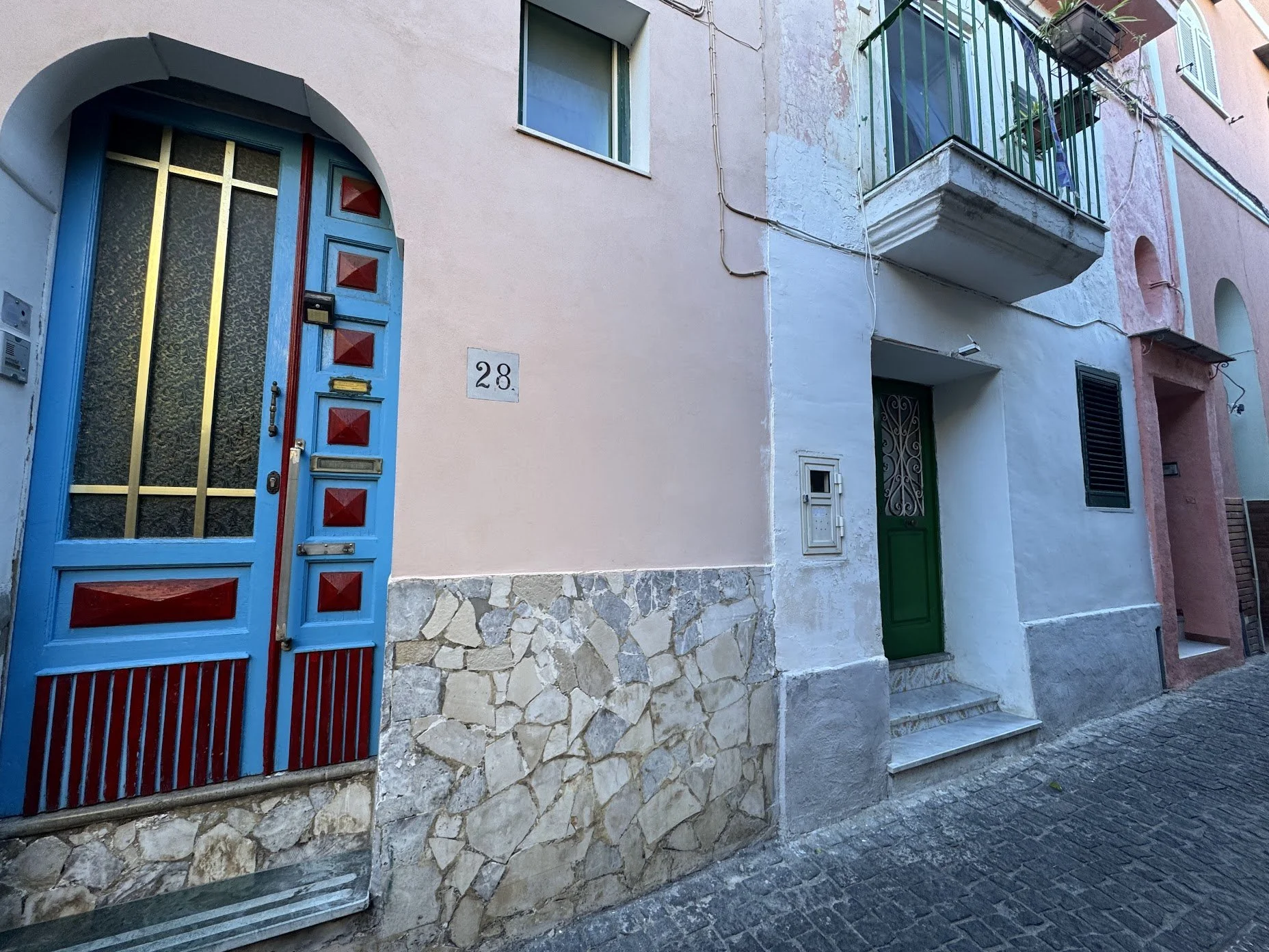 Colorful residential building with a blue front door, a window above, a green door to the right, and a balcony with green railing on the upper level, all set on a cobblestone street.