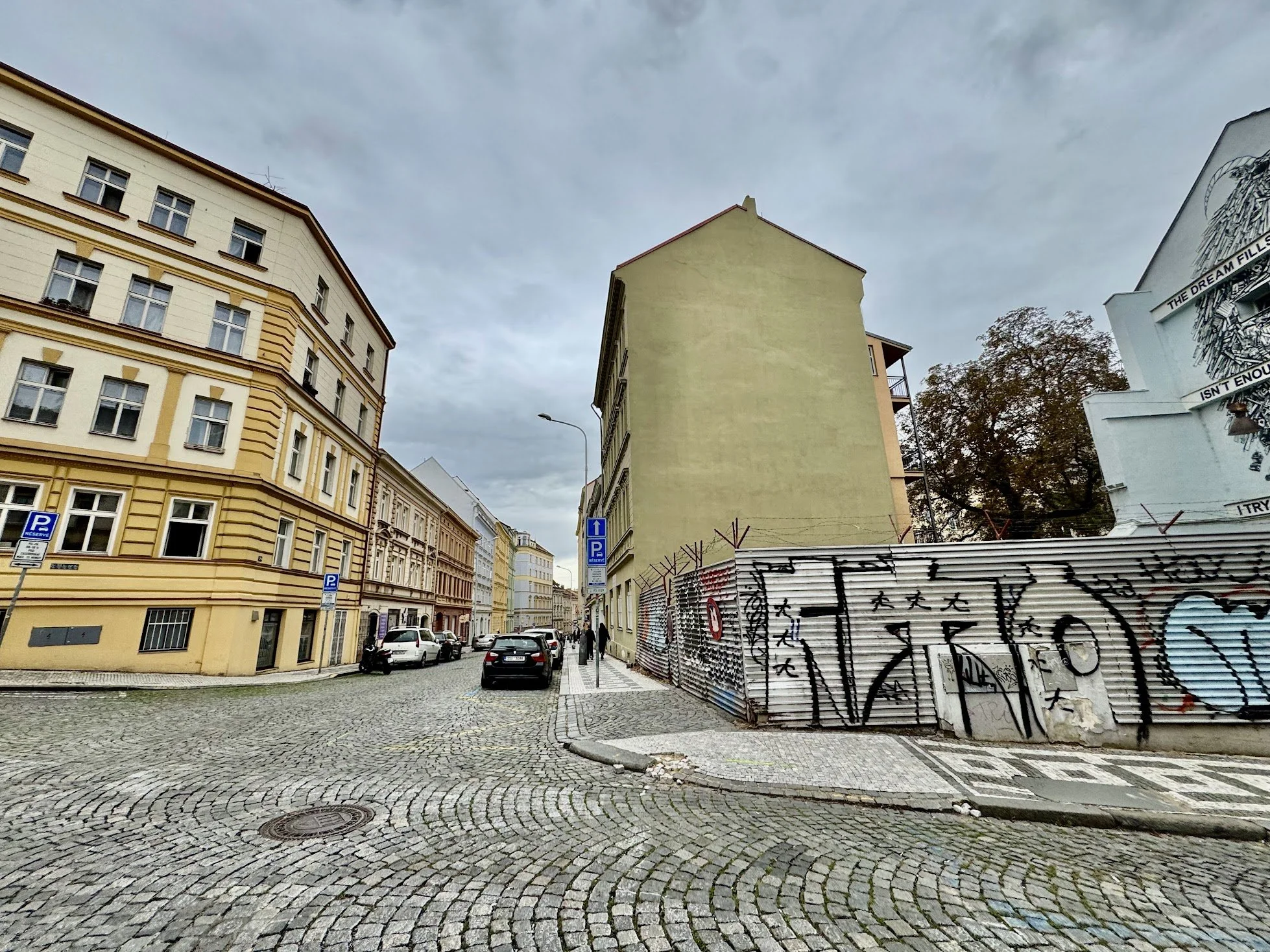 Street scene with cobblestone paving and parked cars on the left, tall pastel-colored buildings on either side, graffiti-covered metal fence on the right, and a cloudy sky above.