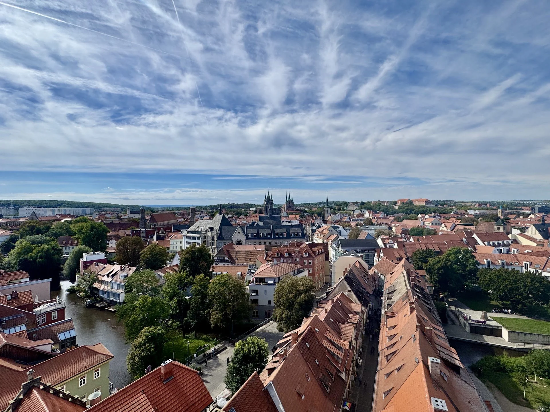 A cityscape with red rooftop buildings, a river, and multiple church spires under a bright blue sky with wispy clouds.