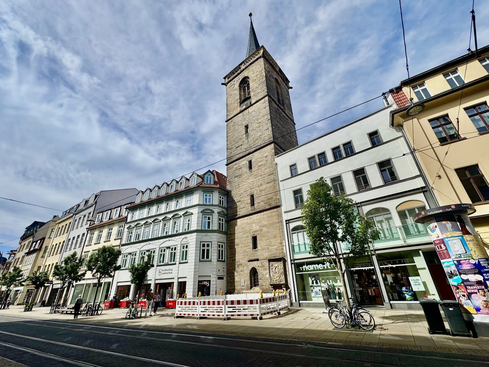 City street scene with historic tower, modern buildings, bicycles, pedestrians, and urban infrastructure under a cloudy sky.
