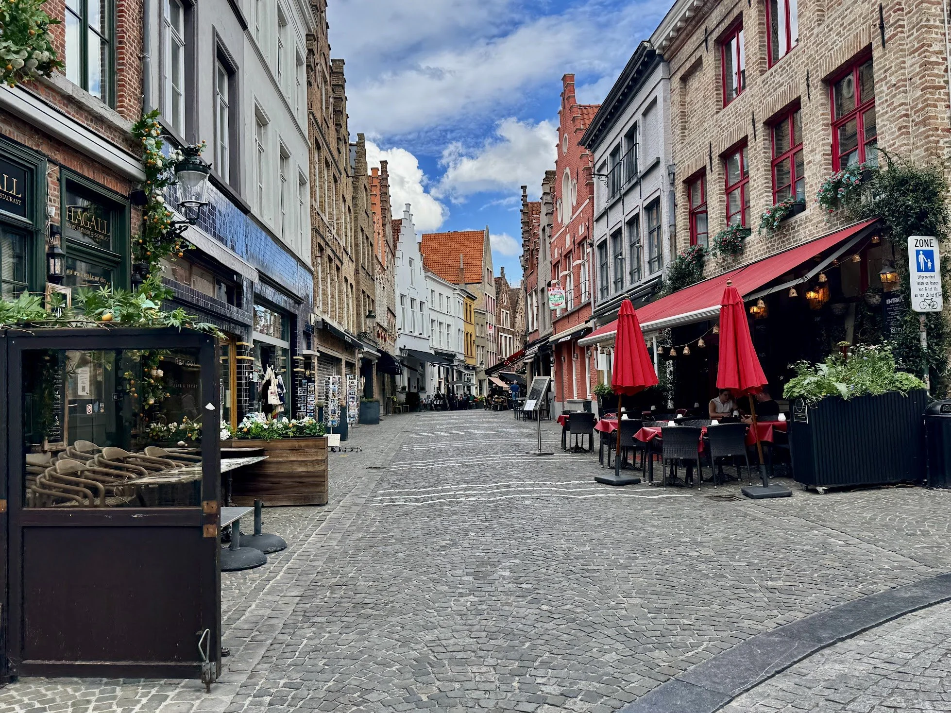 Empty European street with cafes, outdoor seating, colorful historic buildings, and cobblestone pavement on a partly cloudy day.
