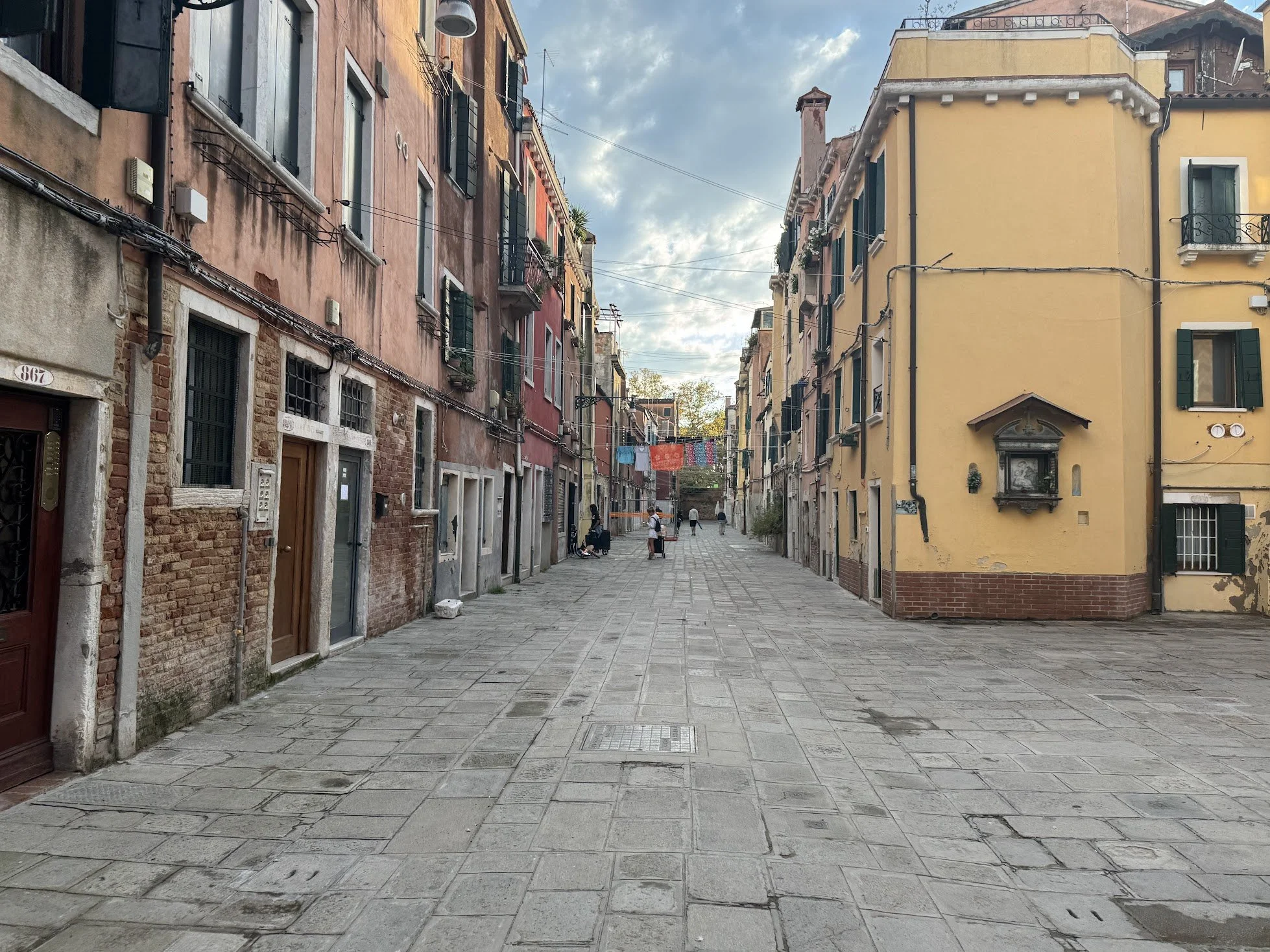 A narrow street in an old European city with colorful buildings on either side, laundry hanging across, and a few people walking.