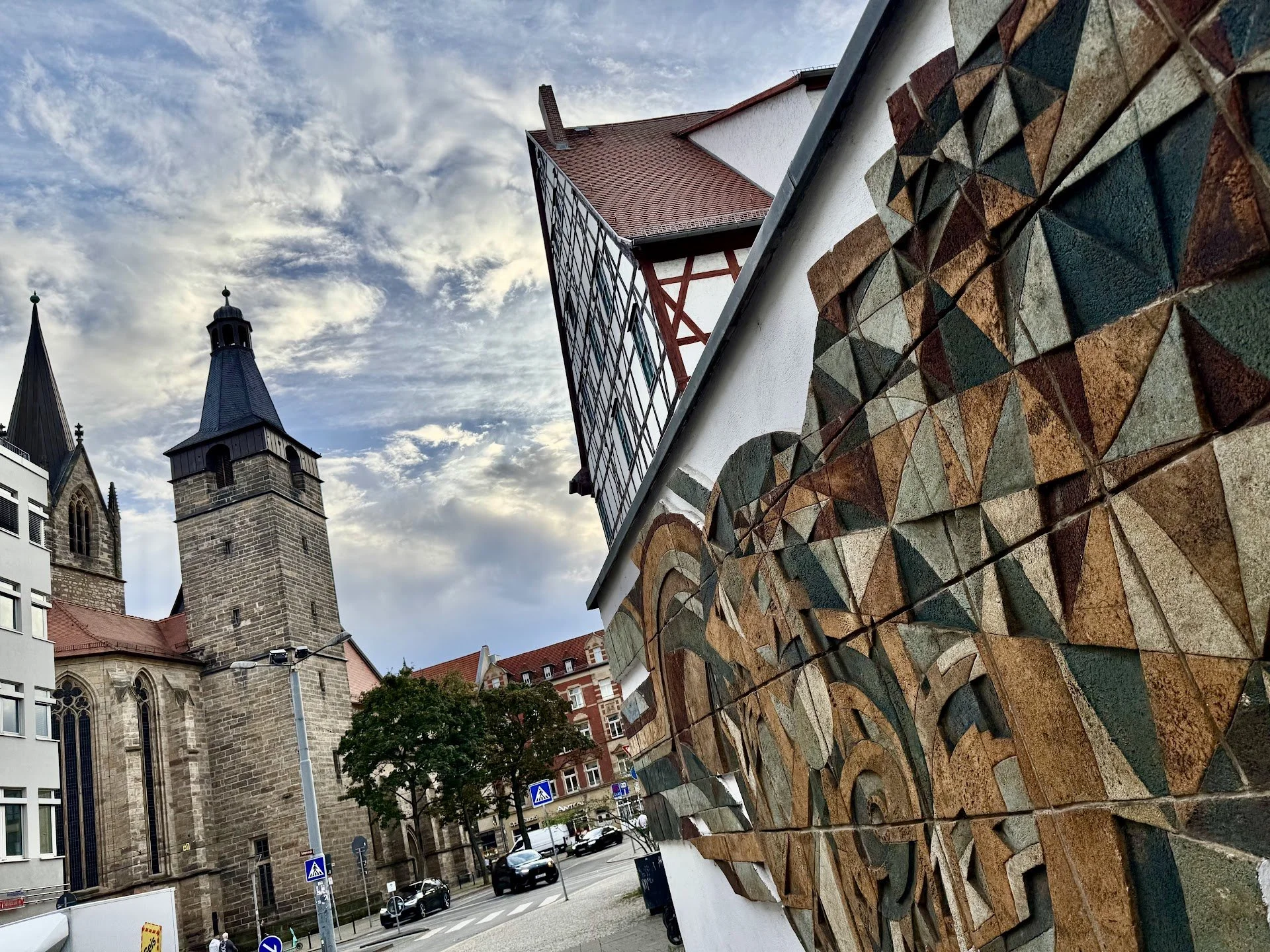 Urban scene featuring stone church tower, modern building with red roof and glass facade, and a wall with colorful stone tile artwork.