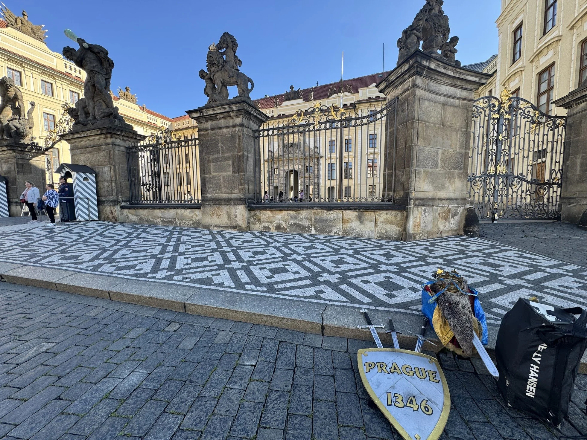 View of a historic gate with ornate black iron gates and four large stone statues of regal figures on pillars. In front, a patterned cobblestone street with pedestrians and a sign that reads 'Prague 1346'.
