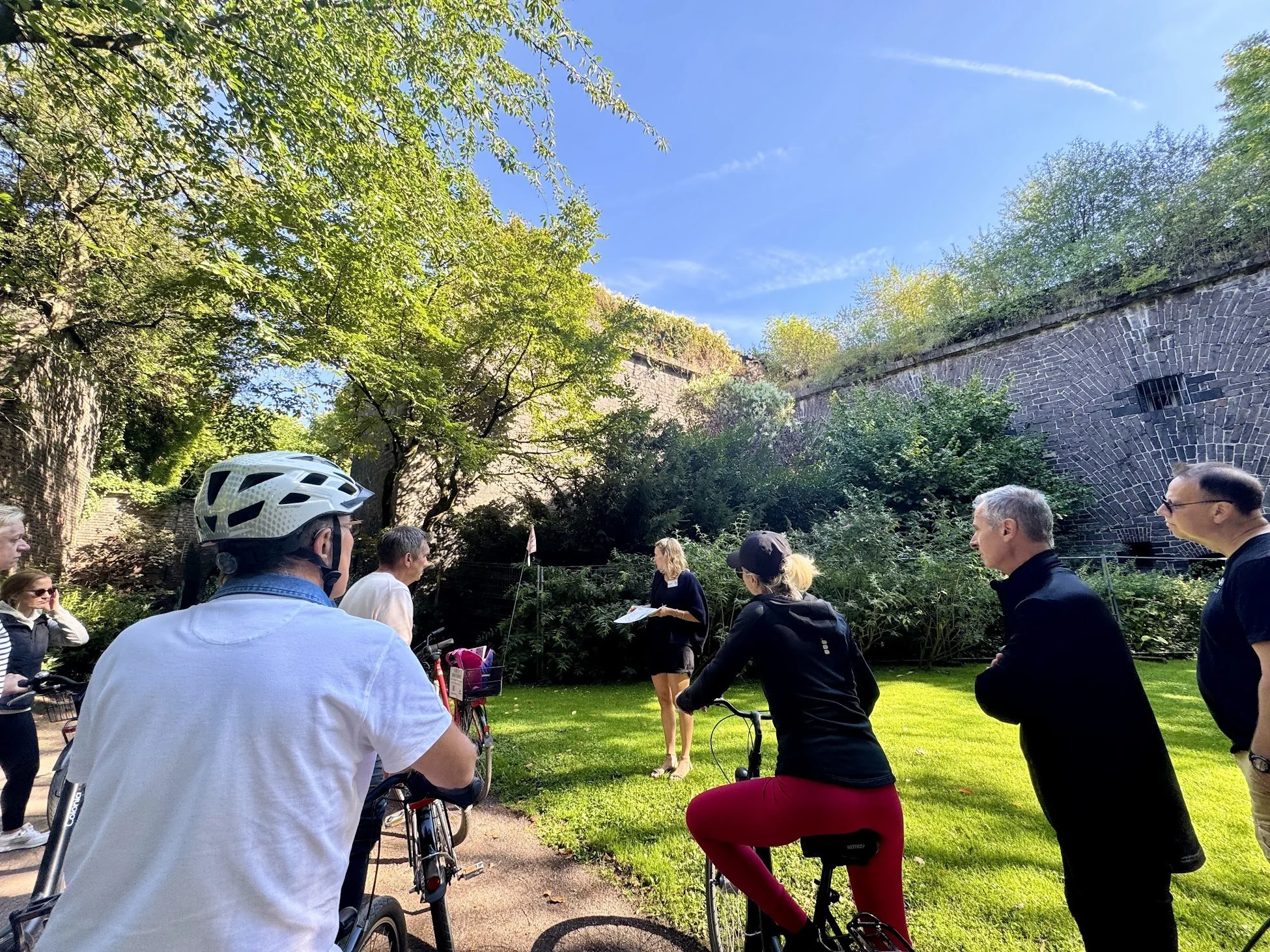 Group of people gathered outdoors in a park, some with bicycles, listening to a woman speaking, with a stone bridge and green trees under a clear blue sky.