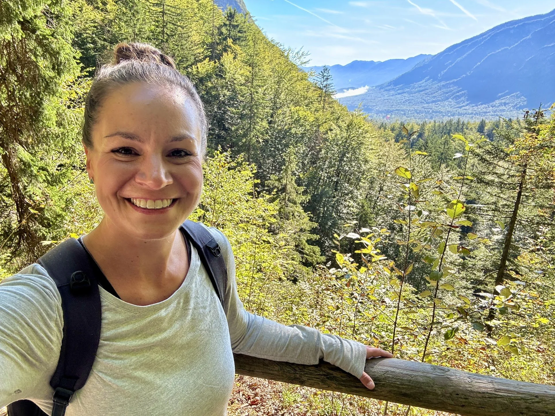 Smiling woman with a backpack taking a selfie in a scenic forest with mountains in the background and a clear blue sky.
