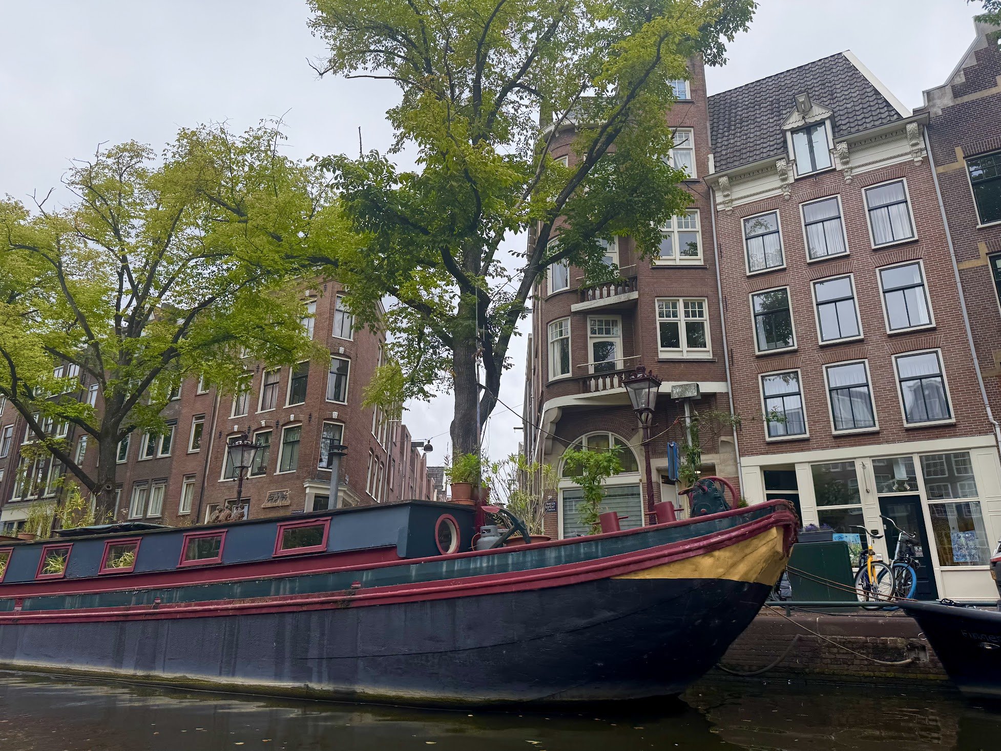 Photo of a canal with a colorful boat in the foreground, tall brick apartment buildings with large windows and decorative facades, trees with green leaves, and a gray sky overhead.