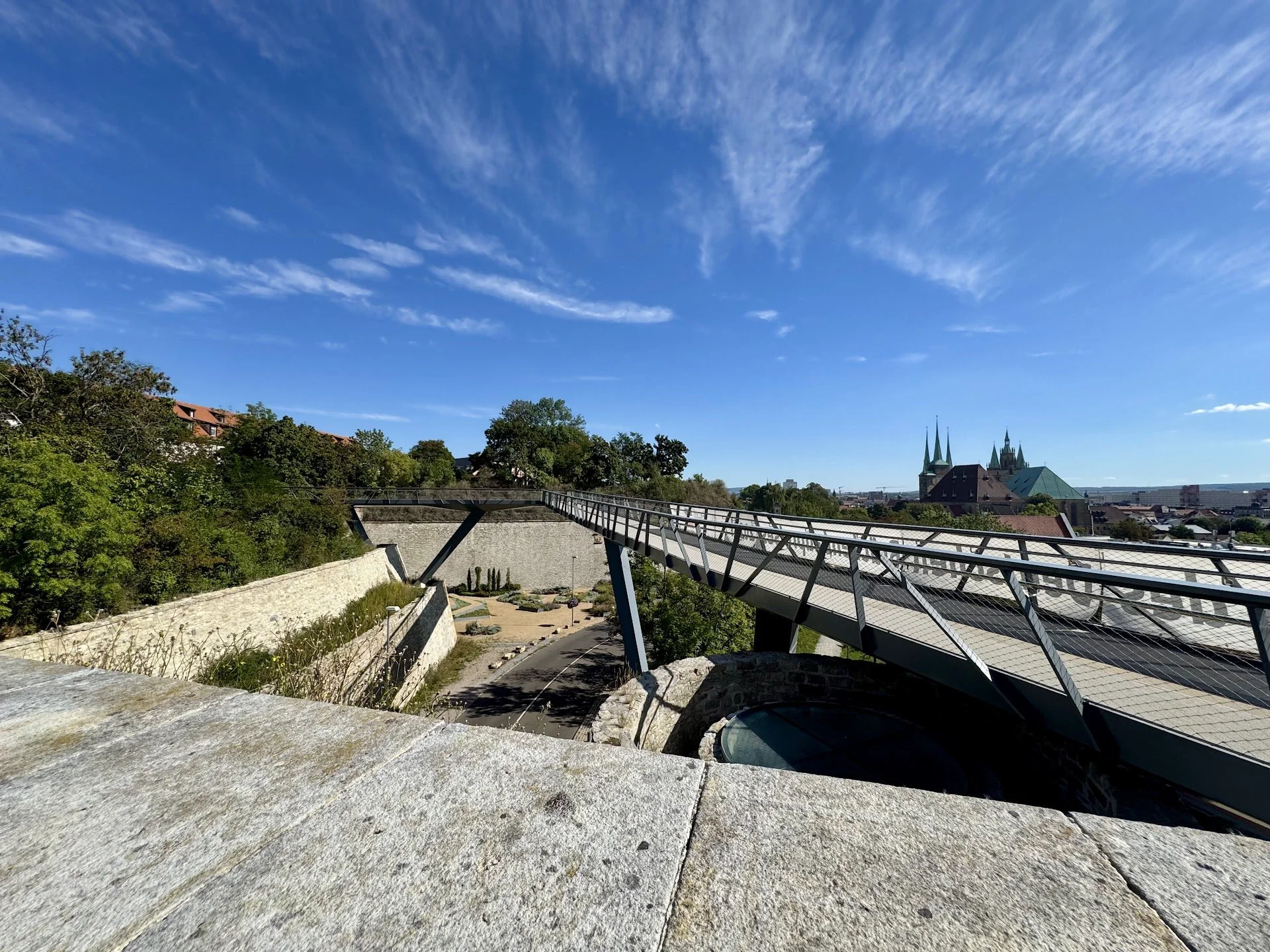 Cityscape view with a modern pedestrian bridge, trees, and historic buildings with church steeples under a blue sky with wispy clouds.