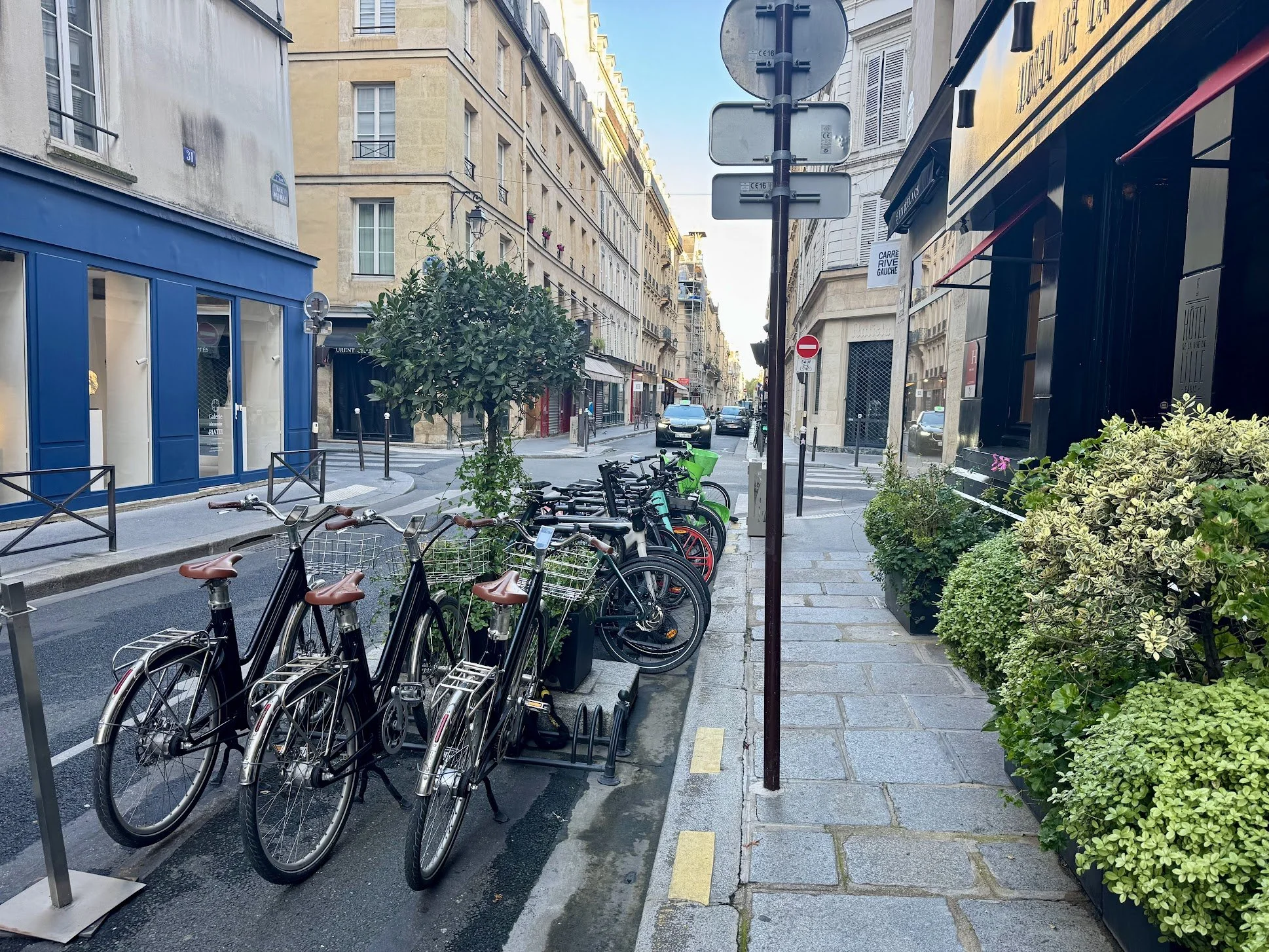 A street in a European city with buildings, parked bicycles, street signs, and green plants.
