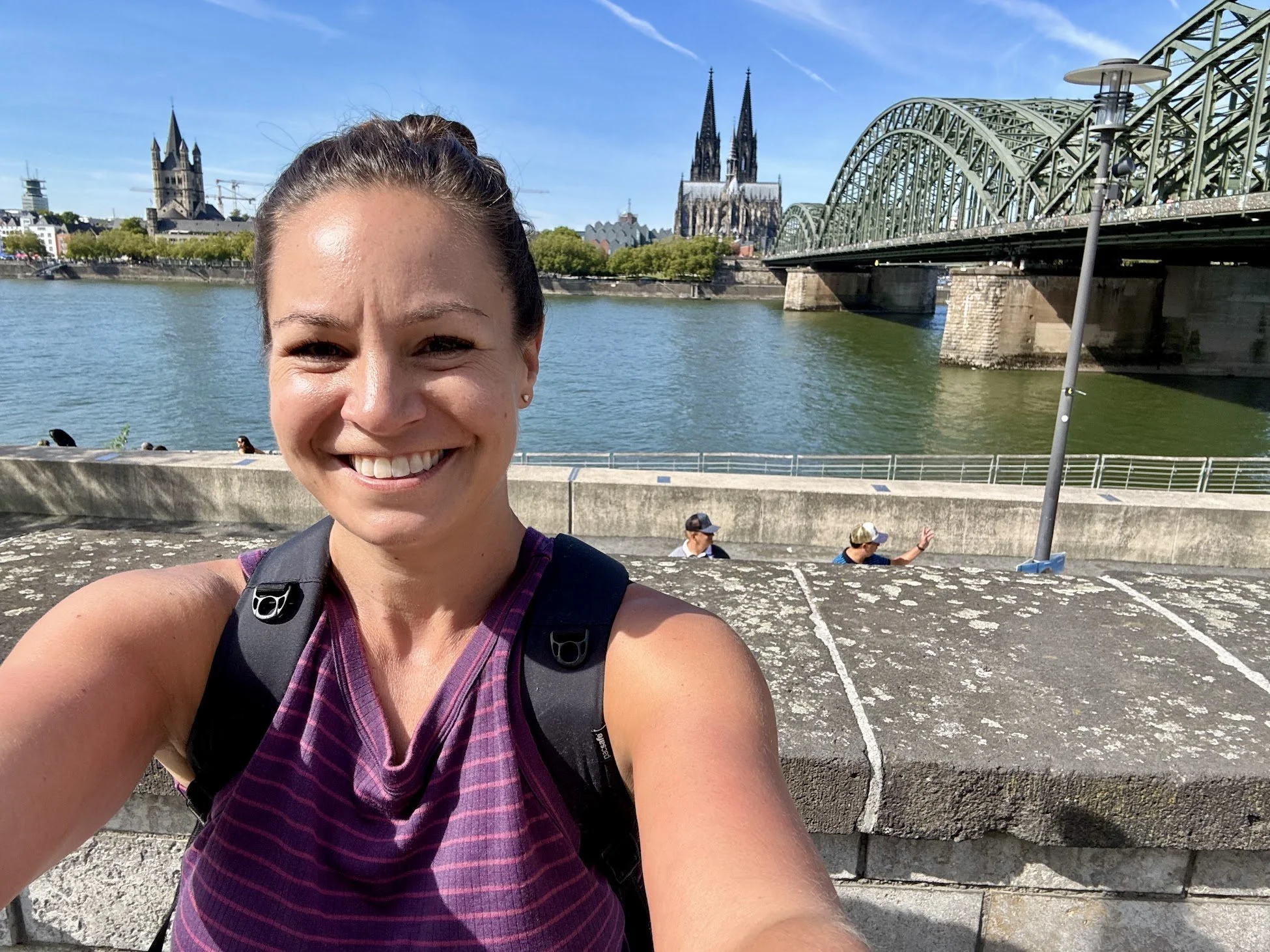 A woman smiling, wearing a purple plaid tank top and a black backpack, taking a selfie near a river with a bridge, a cathedral with twin spires, and a church in the background on a sunny day.