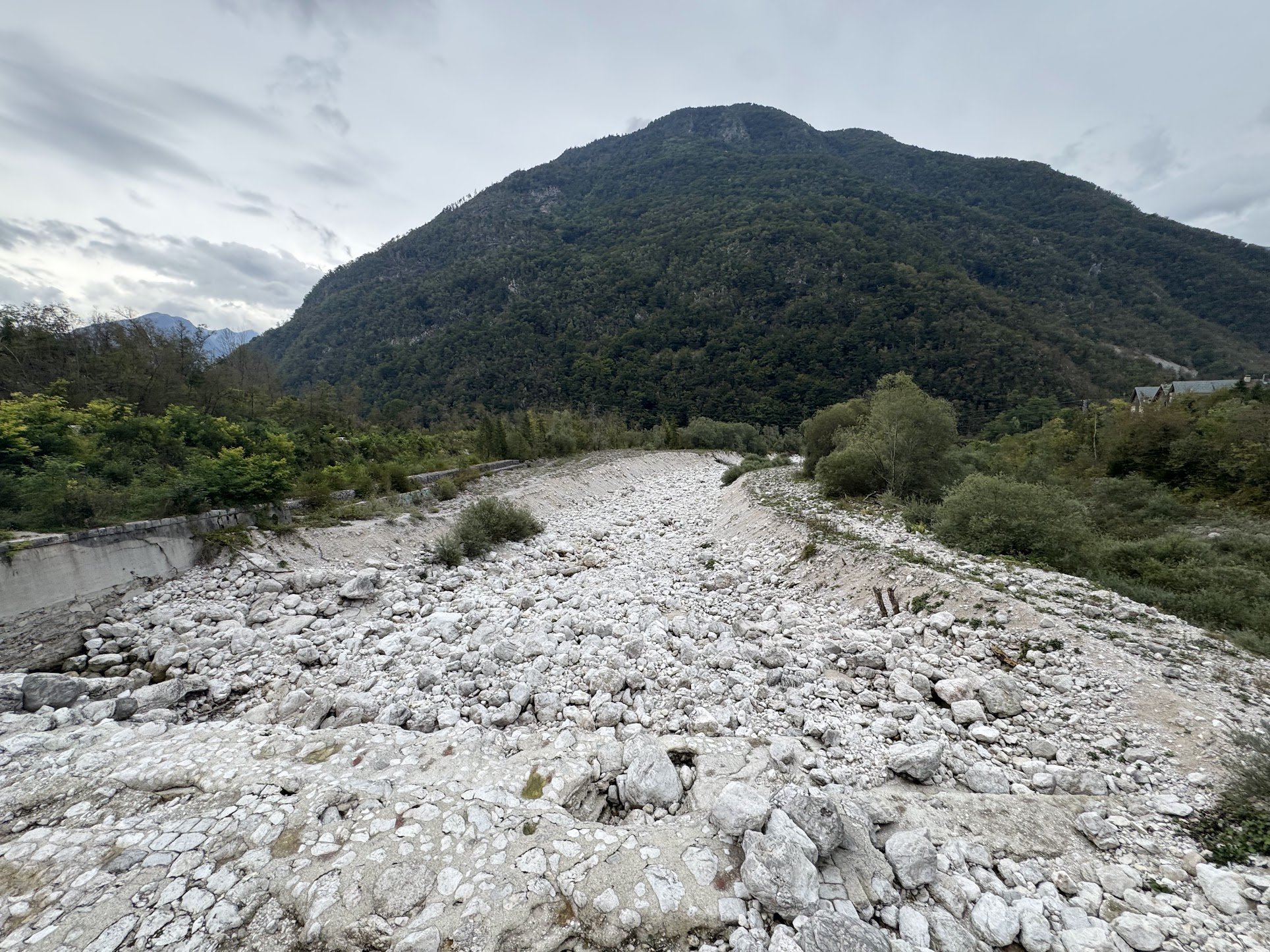 A mountain landscape with a dry rocky riverbed in the foreground, green trees on either side, and a large wooded mountain in the background under a cloudy sky.
