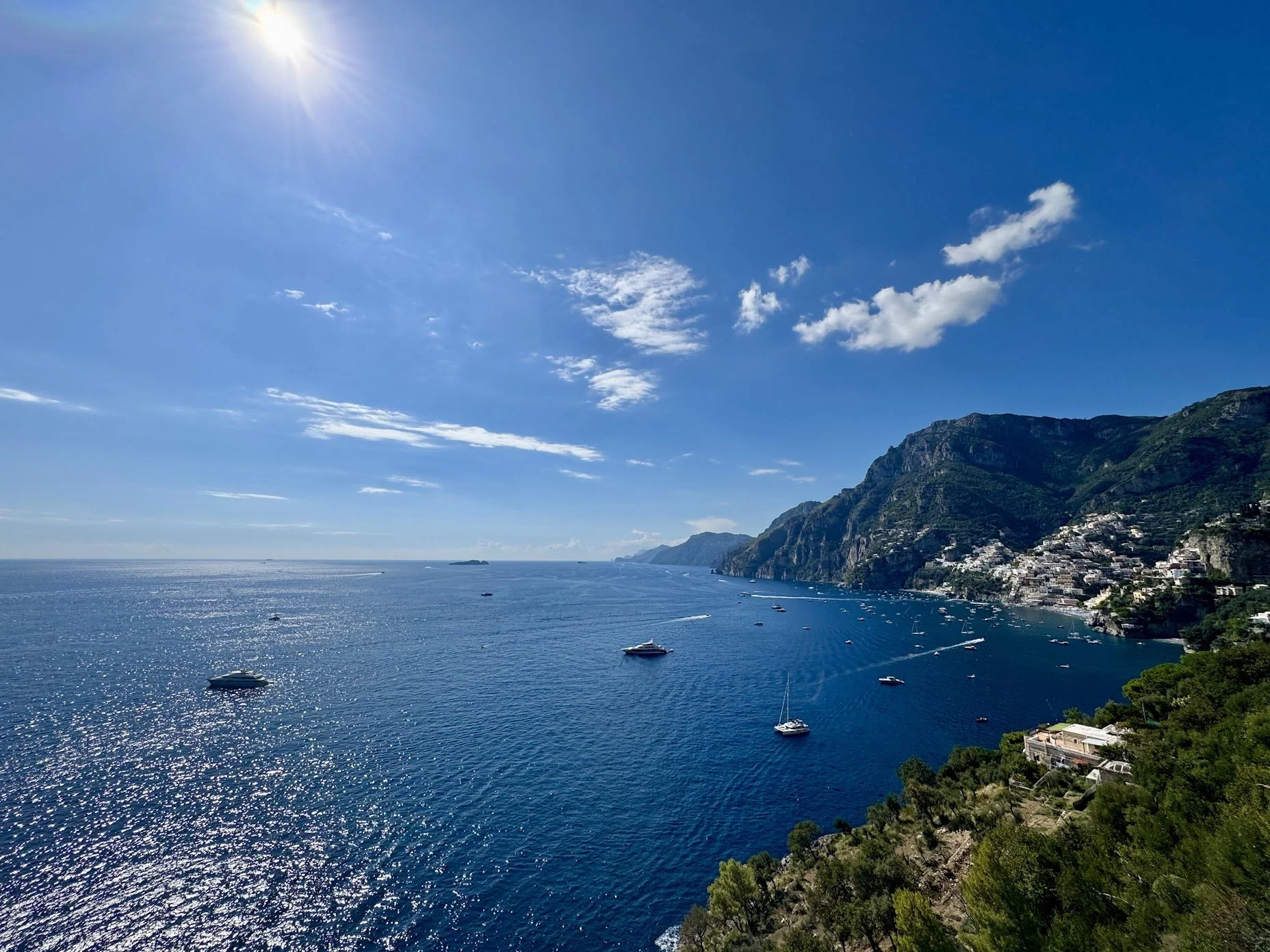 Scenic view of a coastal landscape with blue sea, boats, green hills, and a clear sky with a few clouds and bright sunlight.