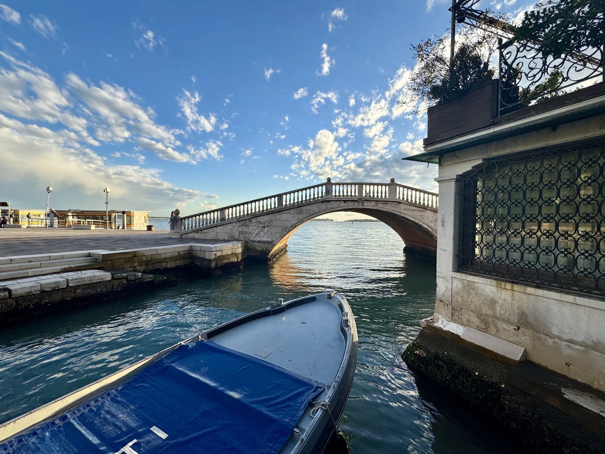 View of a waterfront area with a small boat in the foreground, part of a stone bridge with decorative carvings, a building with ornate black iron railing and a small tree on the balcony, and a sky filled with scattered clouds.