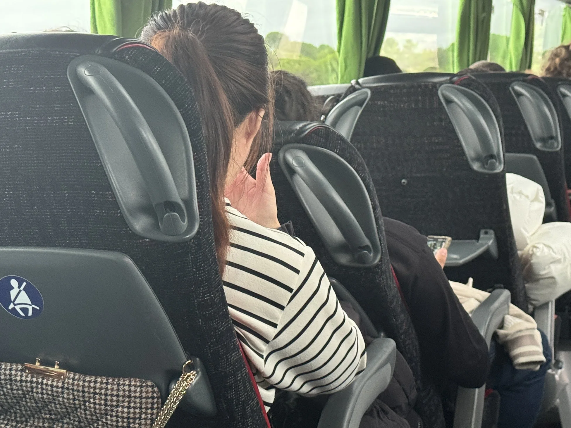 Inside a bus with multiple passengers seated. A woman with long brown hair and glasses is sitting in the middle, wearing a striped shirt, and is using her phone. The seats are black with headrests, and green curtains are visible outside the windows.
