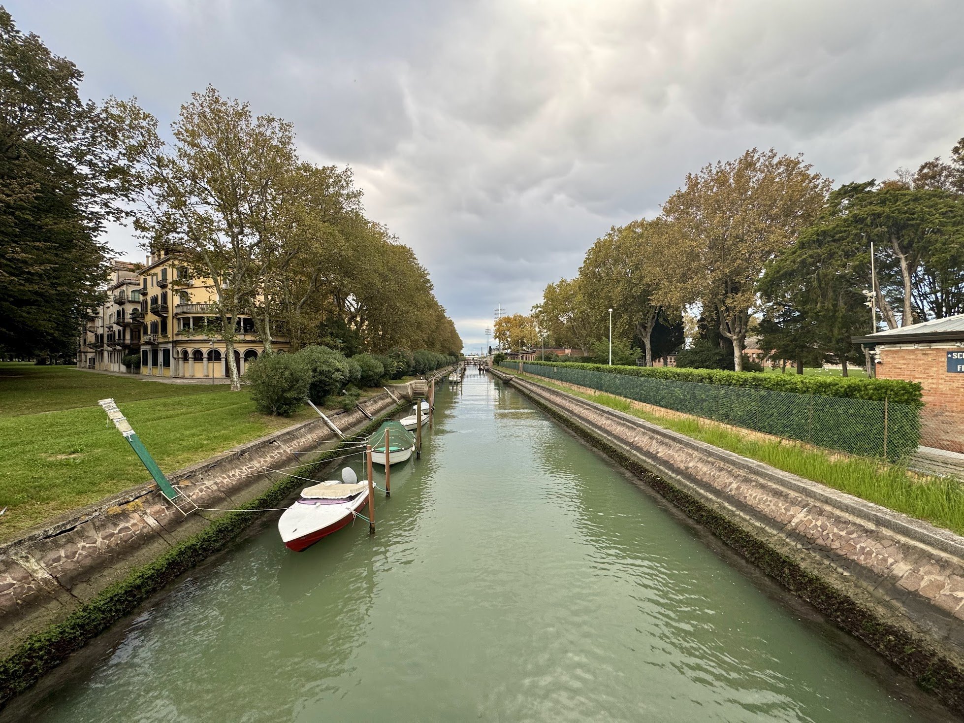 A canal with boats docked along the sides, surrounding a park with trees and a grassy area, under a cloudy sky.