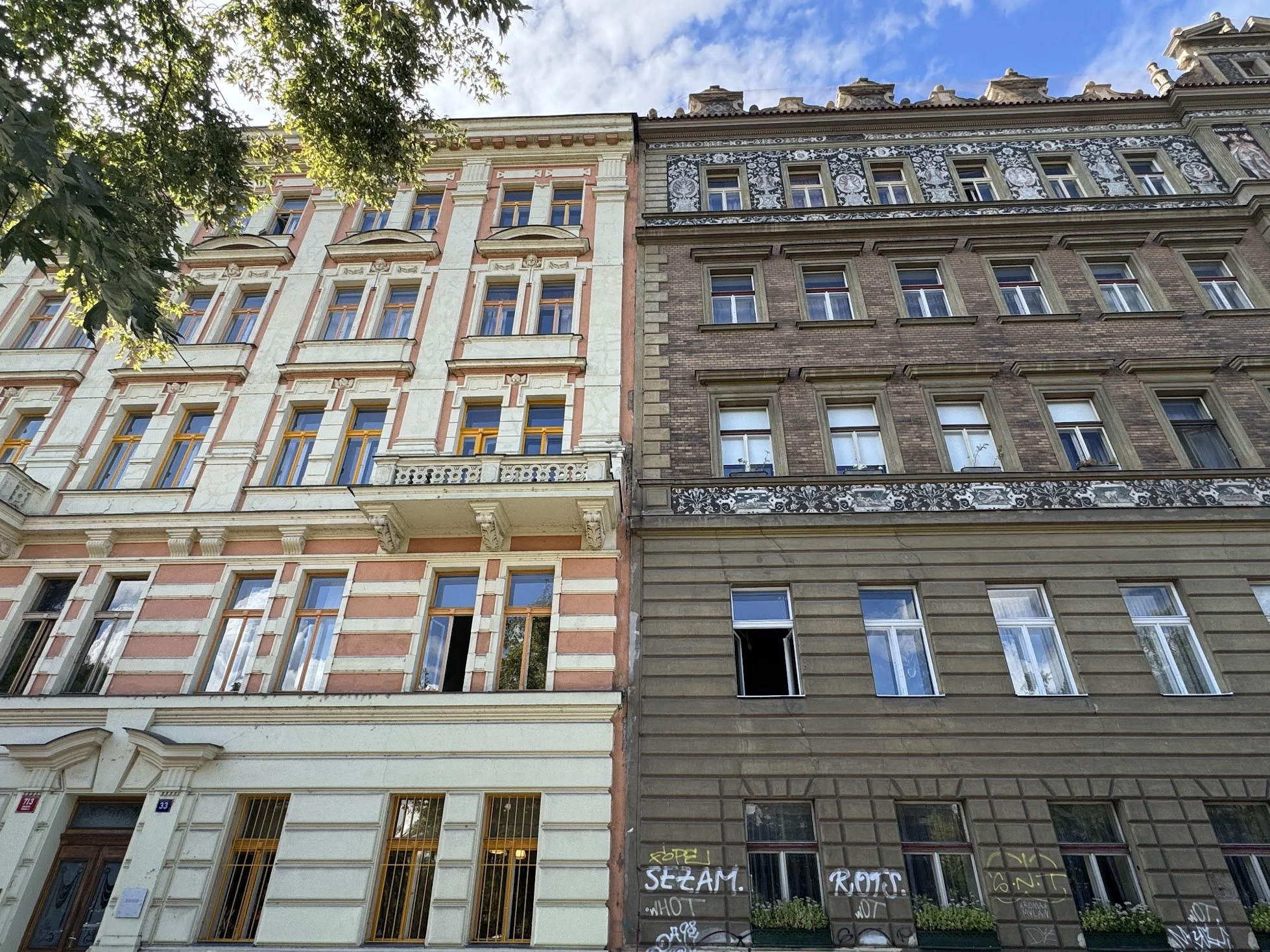 Facade of two adjacent multi-story buildings with ornate window frames, decorative exterior details, and graffiti on the lower part of the right building, under a partly cloudy sky with a tree branch in the upper left corner.