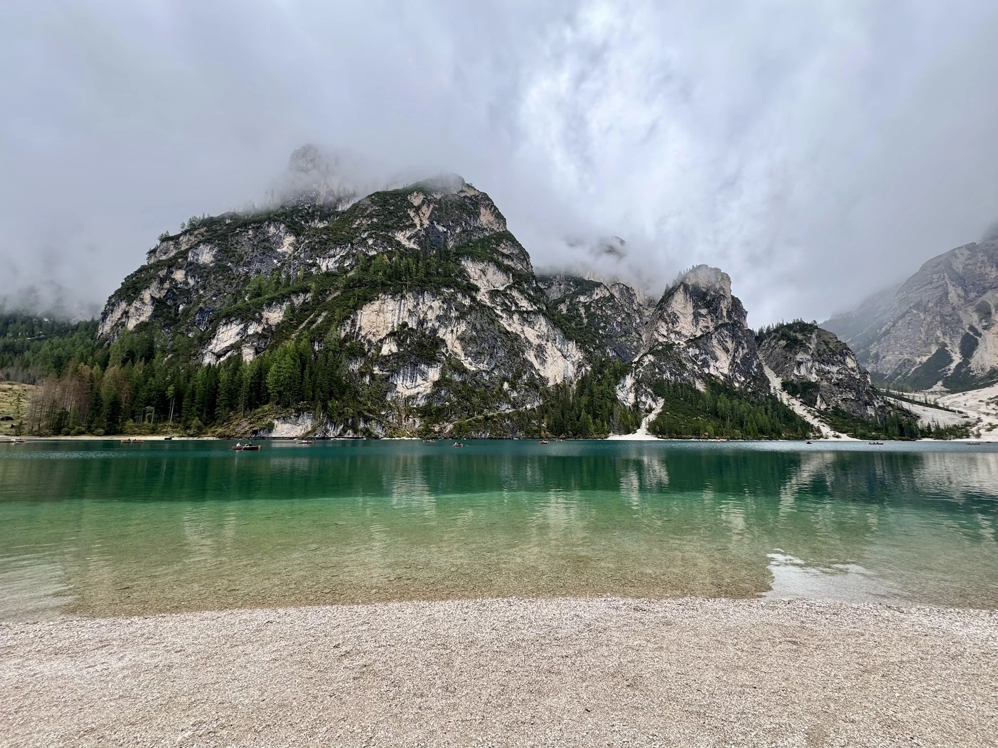 A serene mountain lake scene with a pebble beach, calm turquoise water, forested mountains partially covered in clouds in the background, and a few small boats on the water.