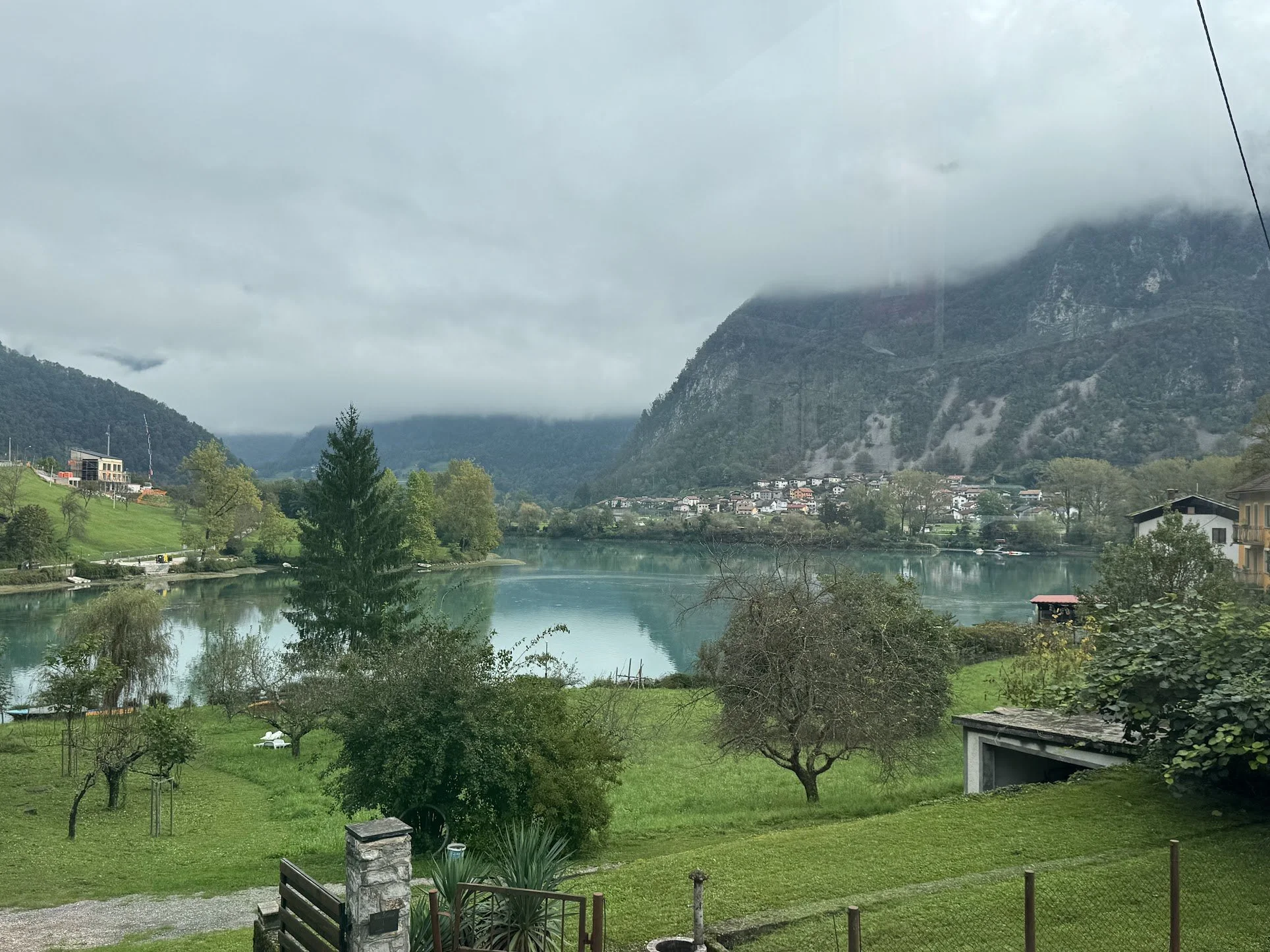 Scenic view of a small town nestled between green hills and a calm river, with fog-covered mountains in the background and a cloudy sky overhead.